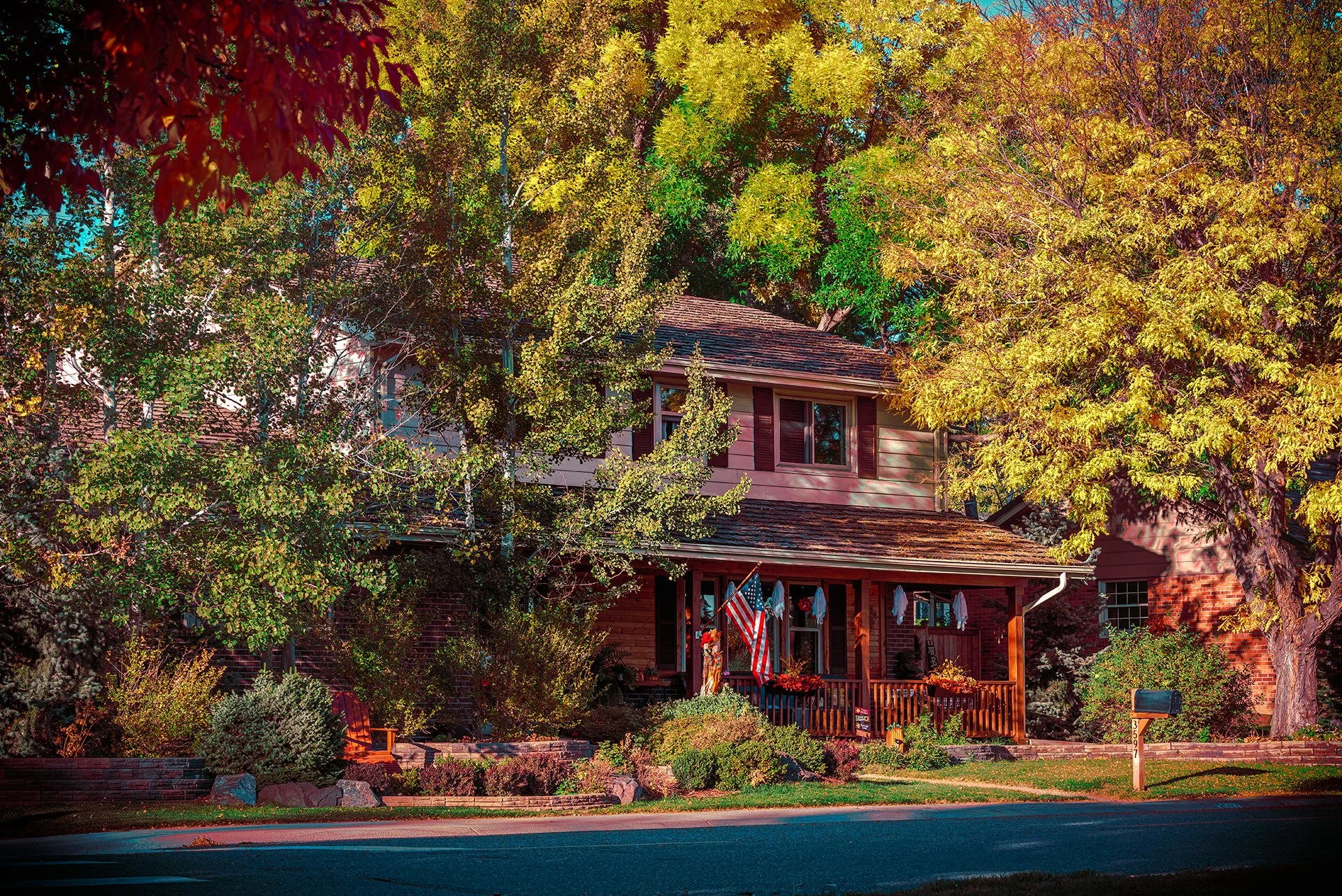 A two-story house with a porch, surrounded by trees with green and yellow leaves, and a well-maintained garden. There is an American flag hanging on the porch.