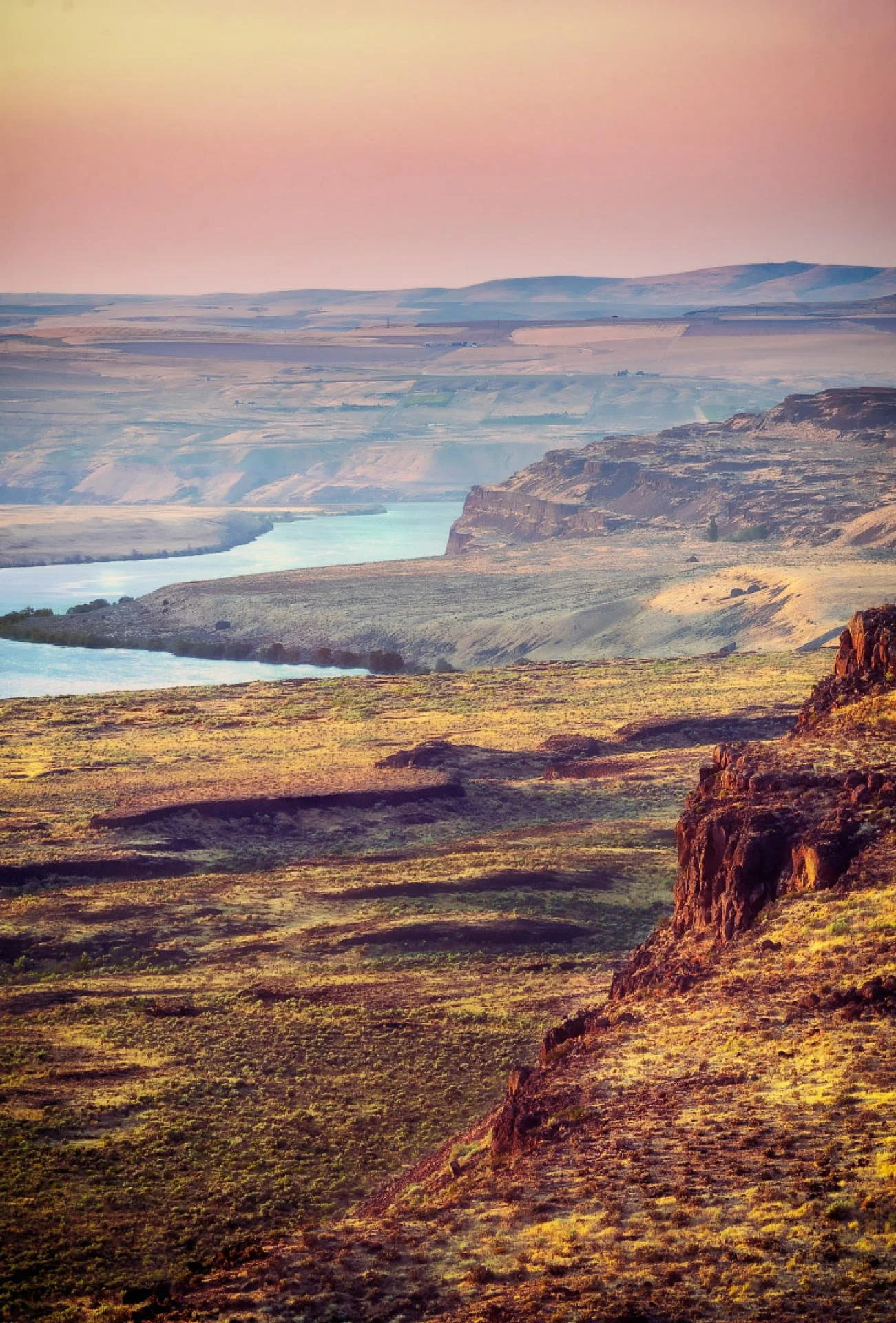 Scenic landscape of a river winding through canyon and rolling hills at sunset with a pink and yellow sky.