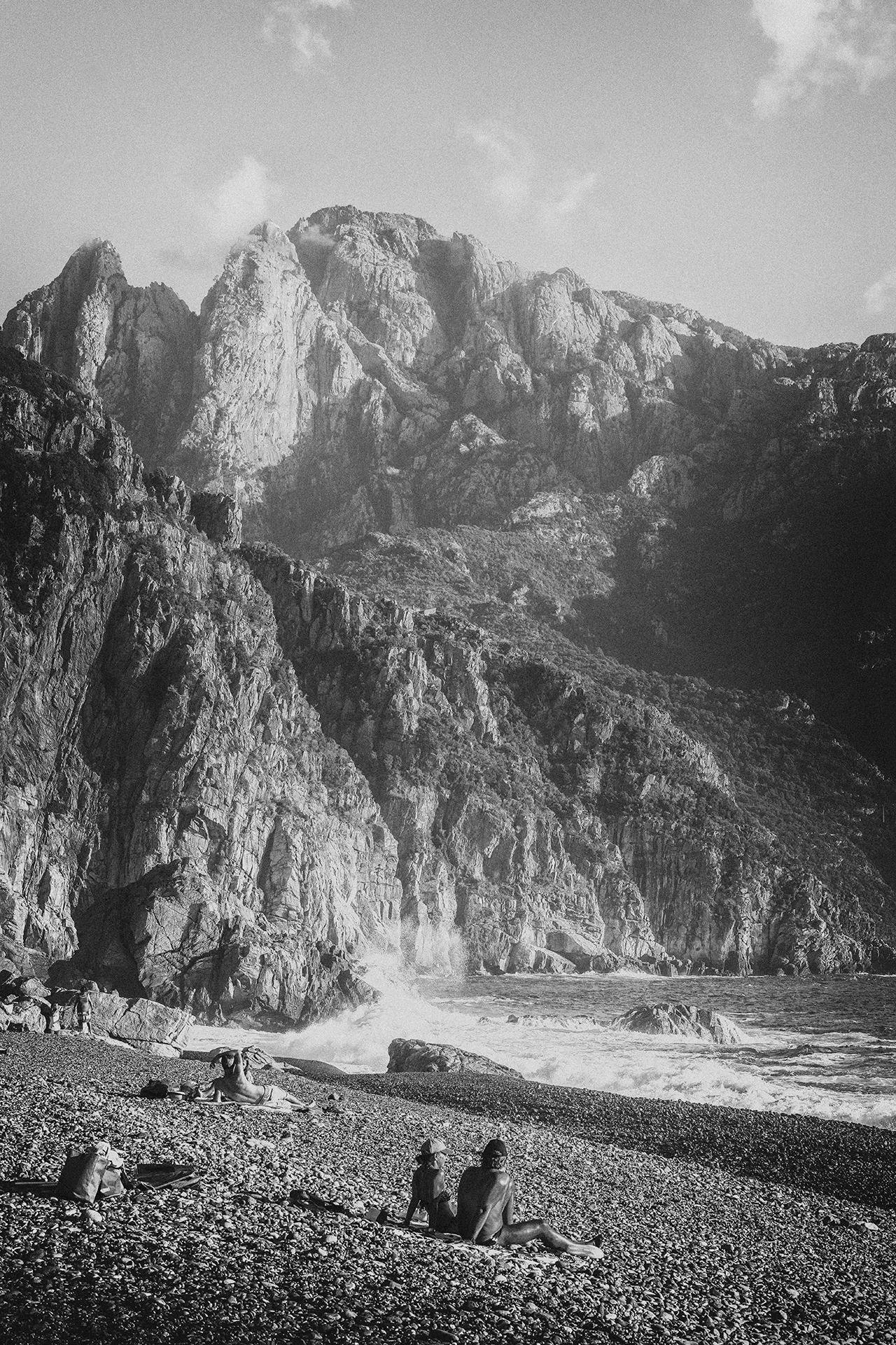 Black and white photo of a rocky beach with steep cliffs and mountains in the background, two people sitting on the beach facing the ocean, one with a hat, and others sunbathing.