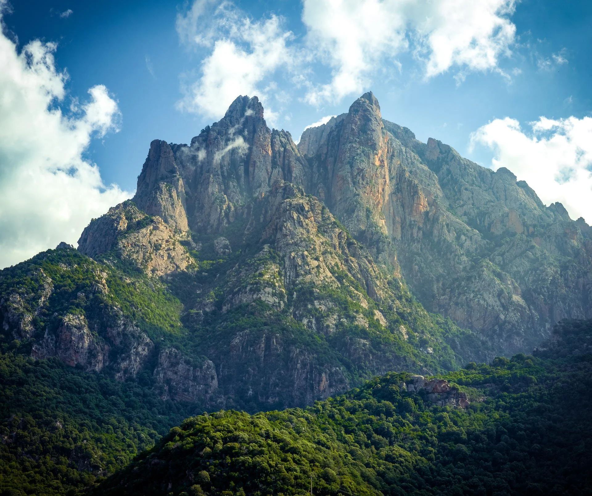 A mountain range with jagged peaks and green forested slopes under a partly cloudy sky.