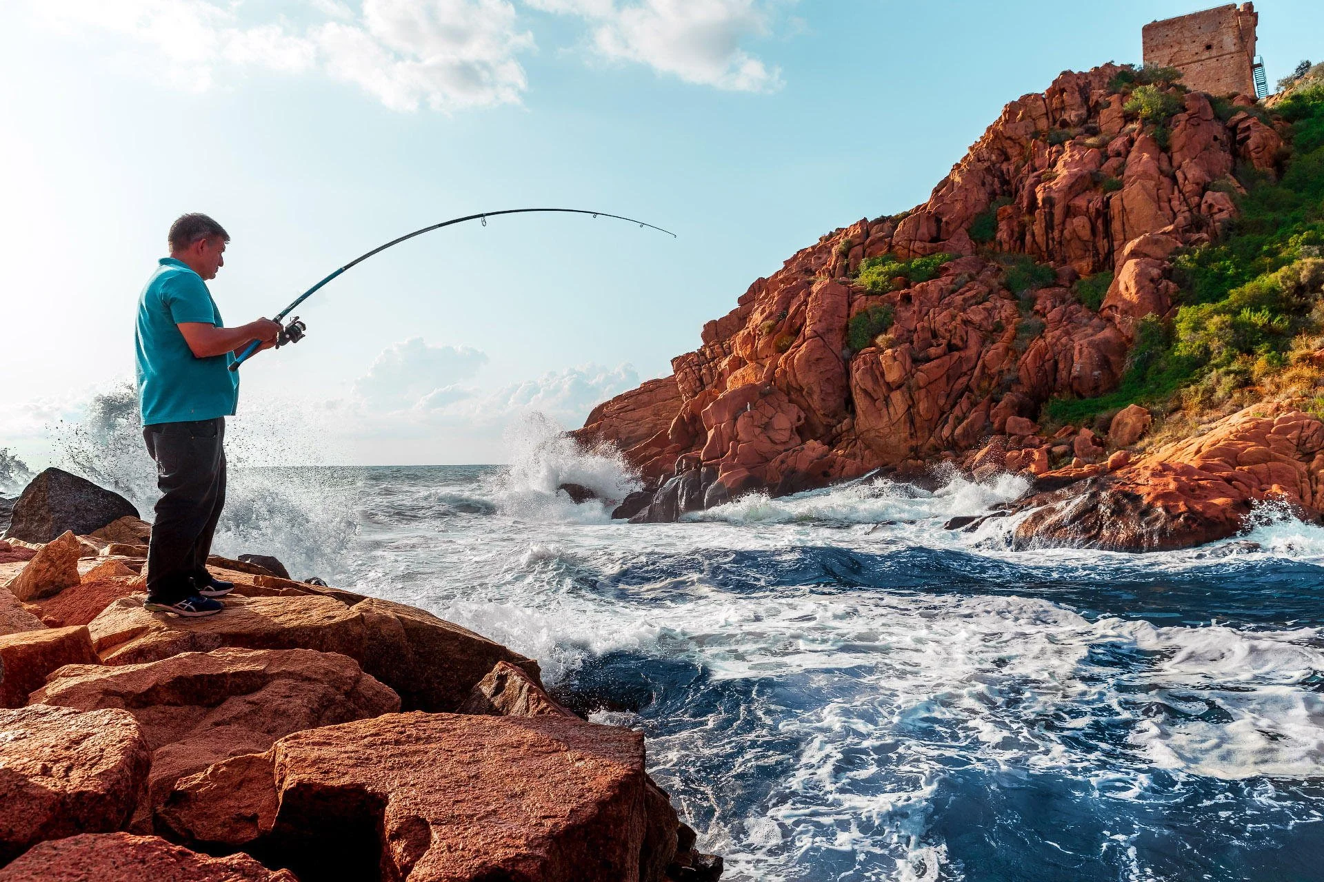A man in a blue shirt and black pants standing on rocks at the shoreline, fishing with a rod as waves crash against the rocks and a red rocky cliff with green vegetation and a structure at the top are in the background.