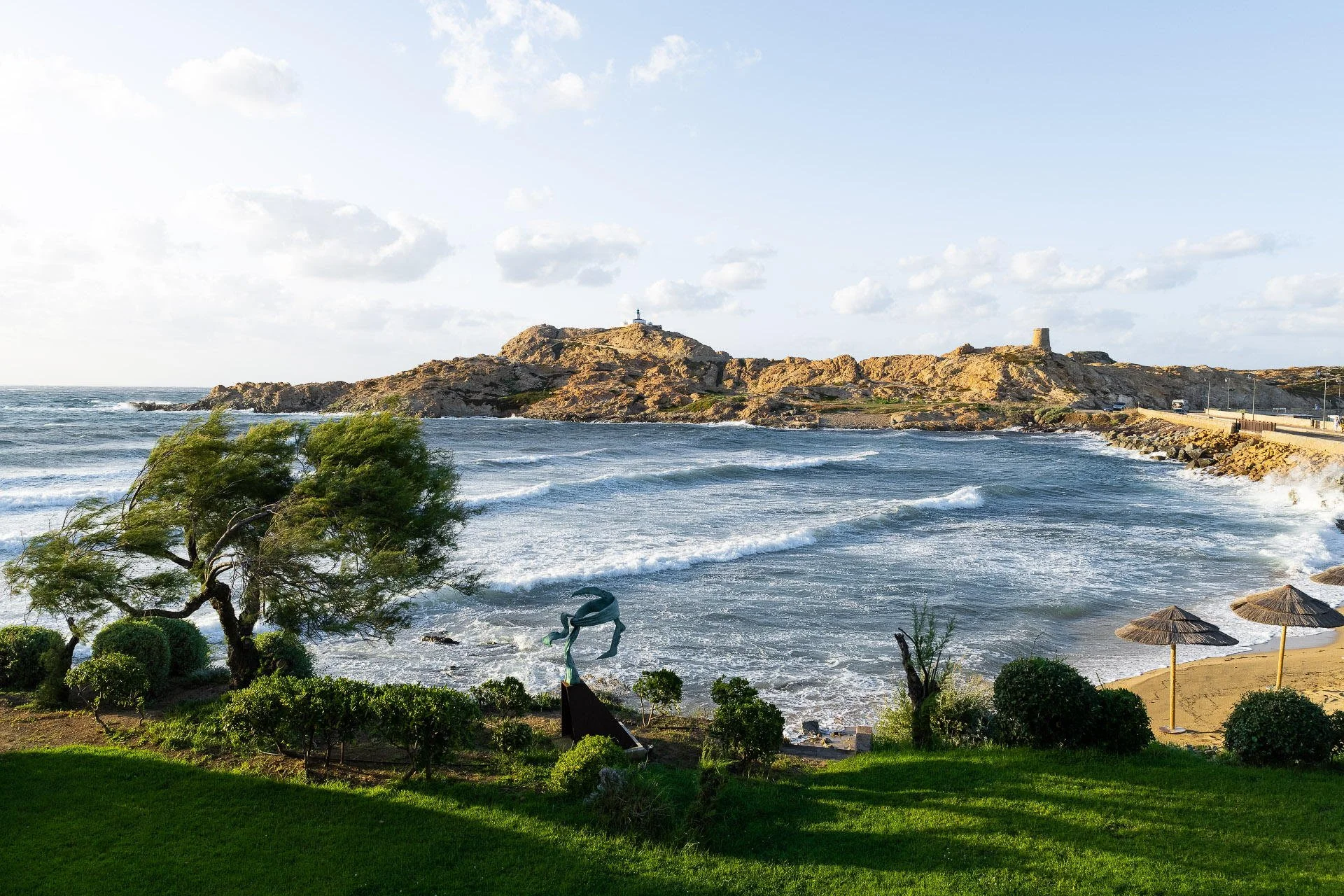 Beach scene with green grass in the foreground, small bushes, a twisted tree, and a metal sculpture of a heron. The sandy beach has two straw umbrellas and is bordered by rocks. Ocean waves crash onto the shore, with a rocky hill in the background to