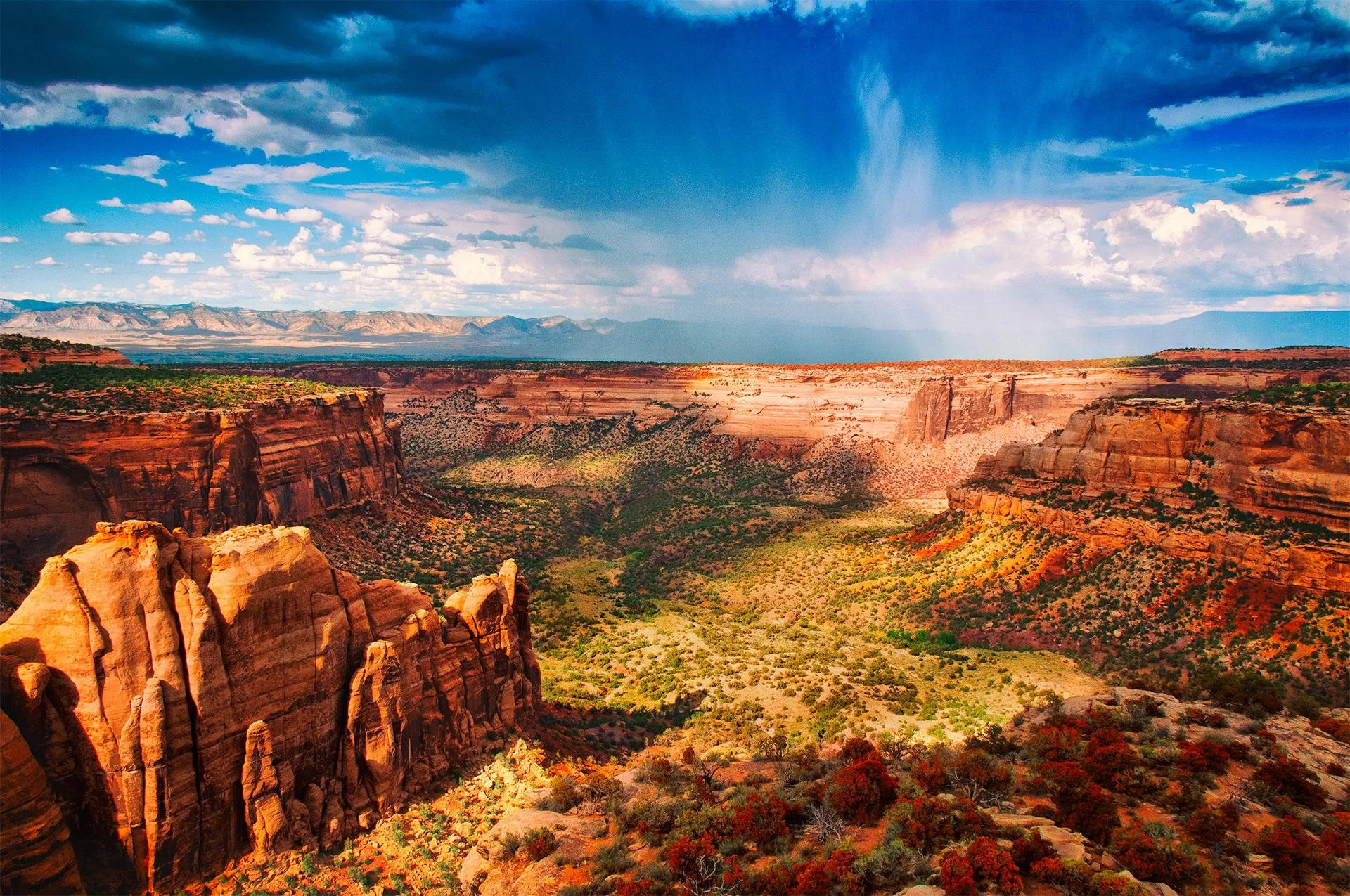 A vast canyon landscape with towering red and orange cliffs, green vegetation in the valley, and a partly cloudy sky with patches of sunlight and rain visible in the distance.