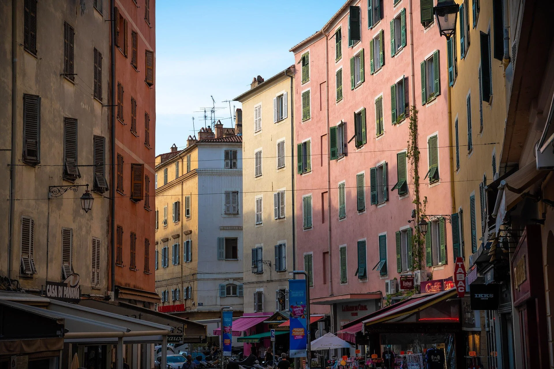 Colorful buildings with shuttered windows in a European city street, with outdoor cafes and shops.