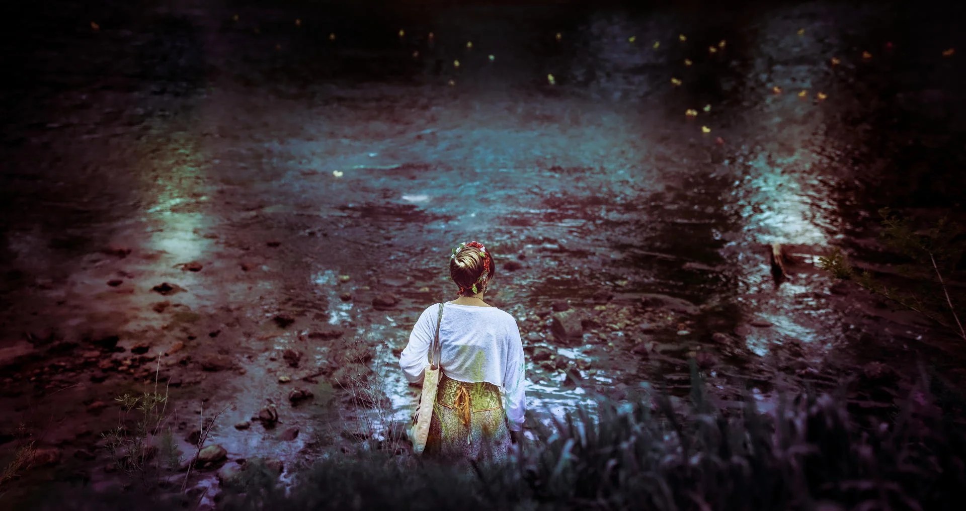 A person in a gray shirt and a flower crown stands at the edge of a body of water at night, with colorful reflections on the surface.