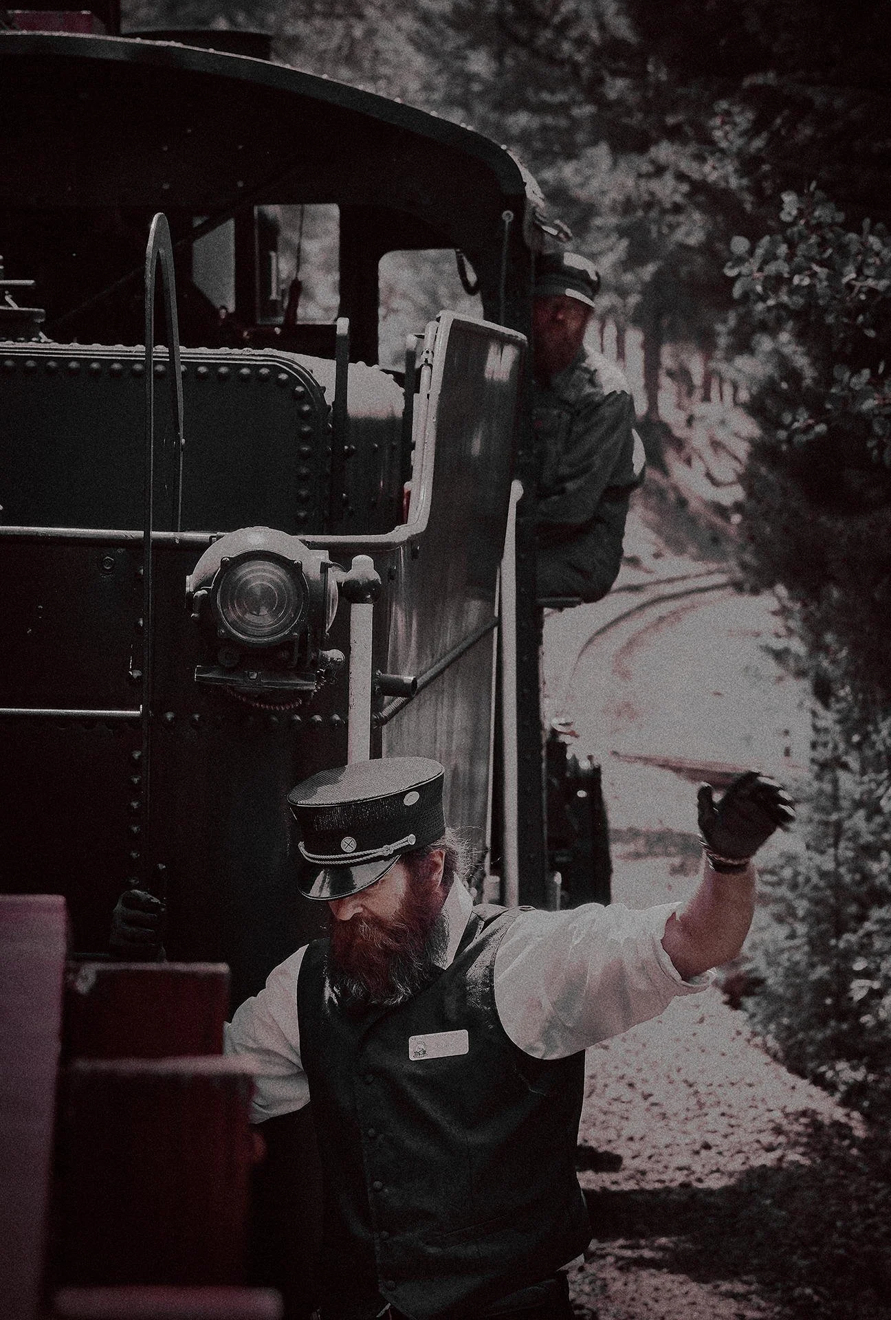 Two men dressed in vintage train conductor uniforms working on or inspecting a steam locomotive on a railroad track surrounded by trees.