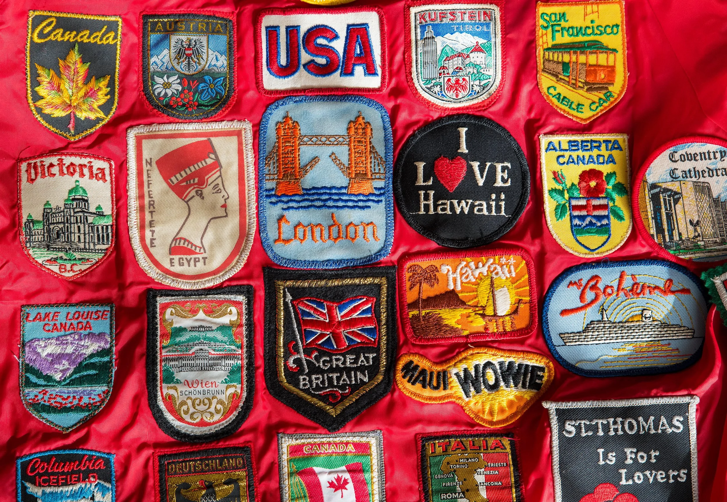 A collection of colorful fabric patches representing various countries and locations, including Canada, Austria, USA, San Francisco, Hawaii, London, Alberta, Egypt, Great Britain, and others, arranged on a red background.