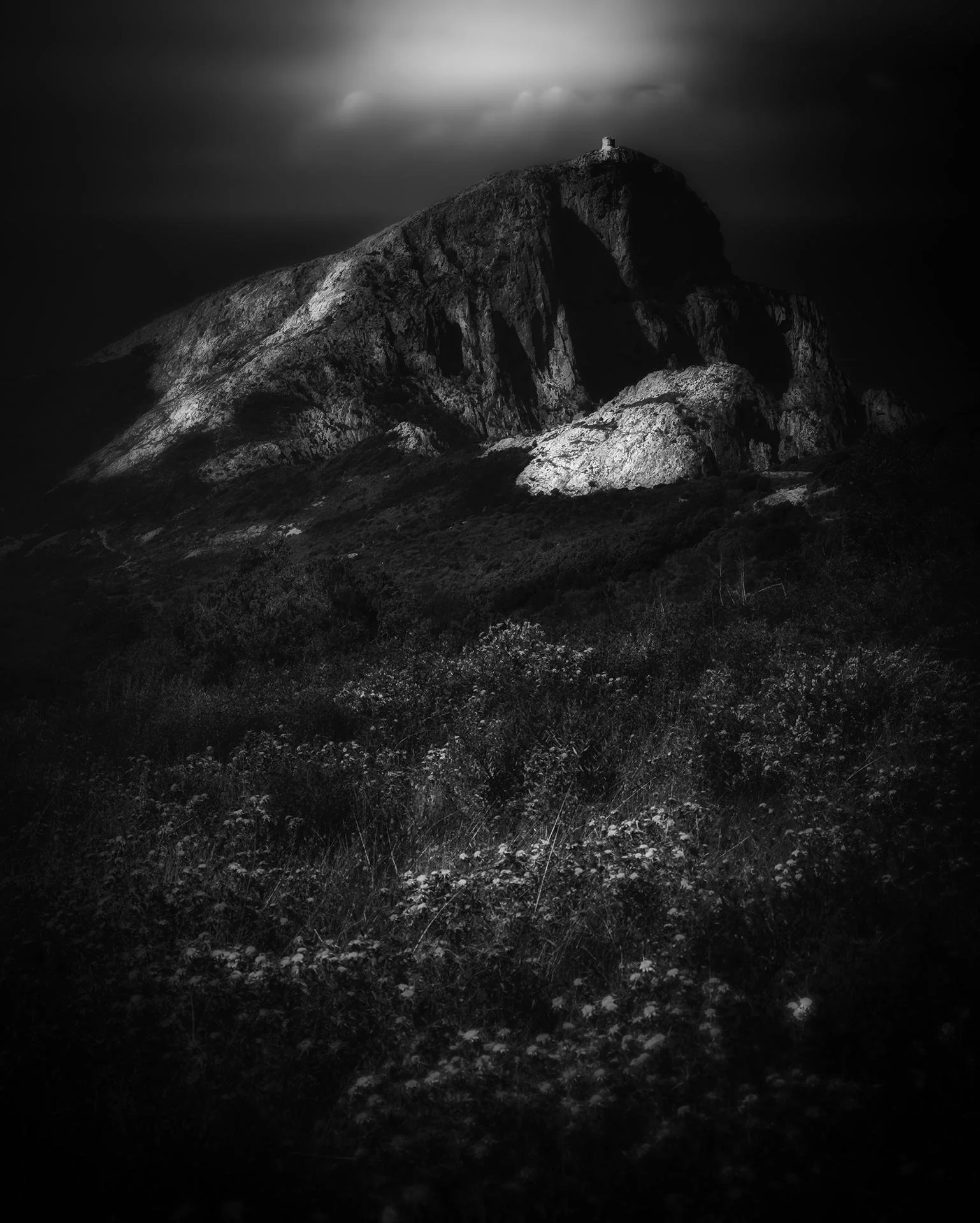 A dark, moody black and white photograph of a rugged mountain with a small structure at its peak, with cloudy skies overhead.