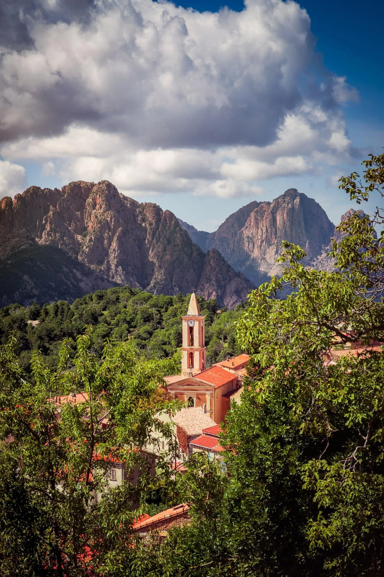A small village surrounded by lush green trees with a church steeple in the center, set against a backdrop of tall, rugged mountain peaks under a partly cloudy sky.