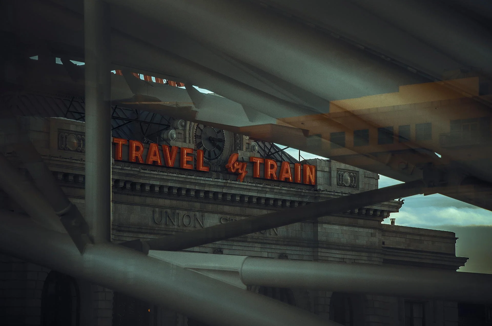 The exterior of a train station with a clock and a sign that reads 'Travel by Train', partially obscured by the reflection of an indoor area.