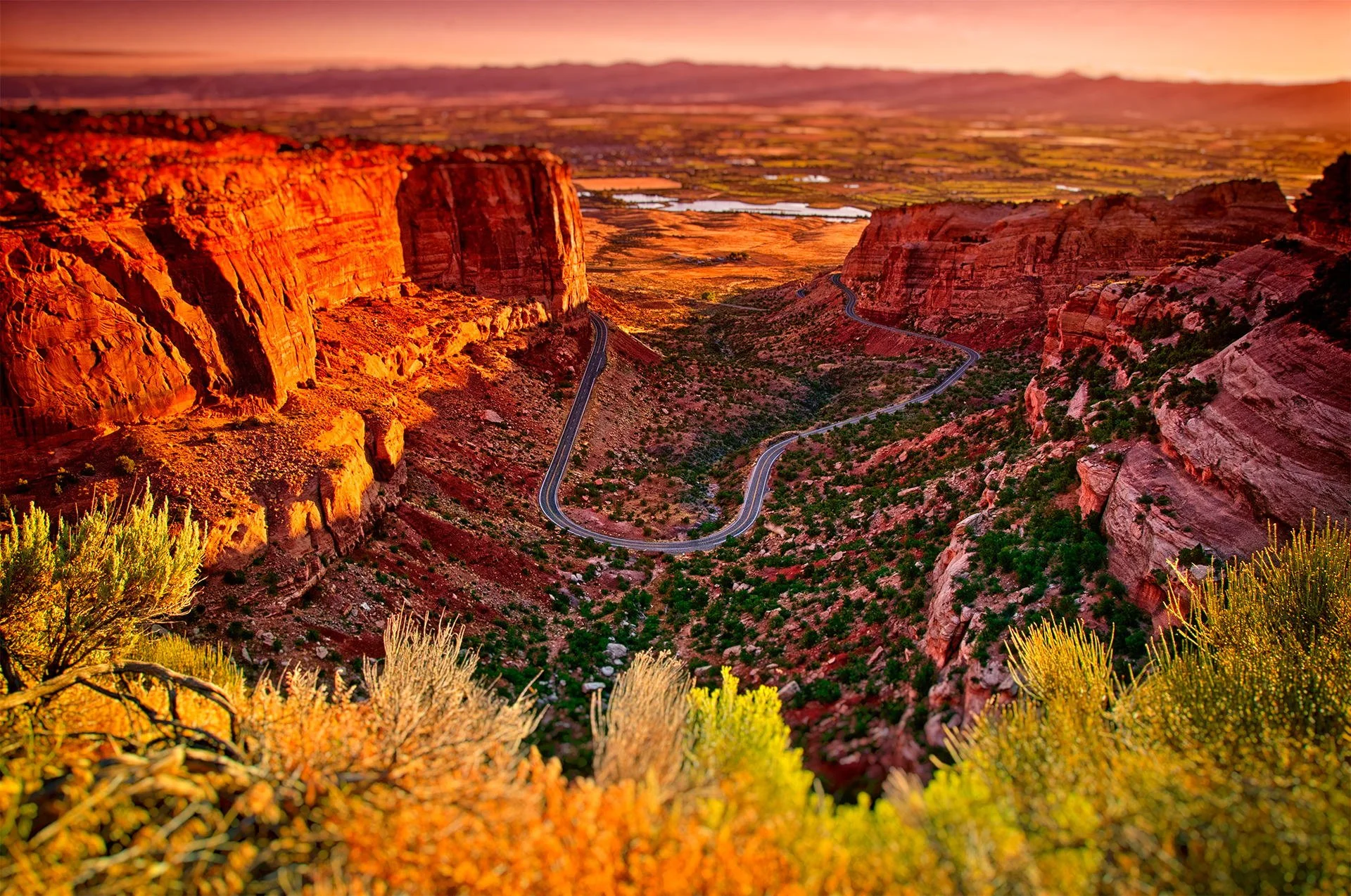 A winding road through a desert canyon with red rock formations, sparse vegetation, and a distant body of water at sunset.