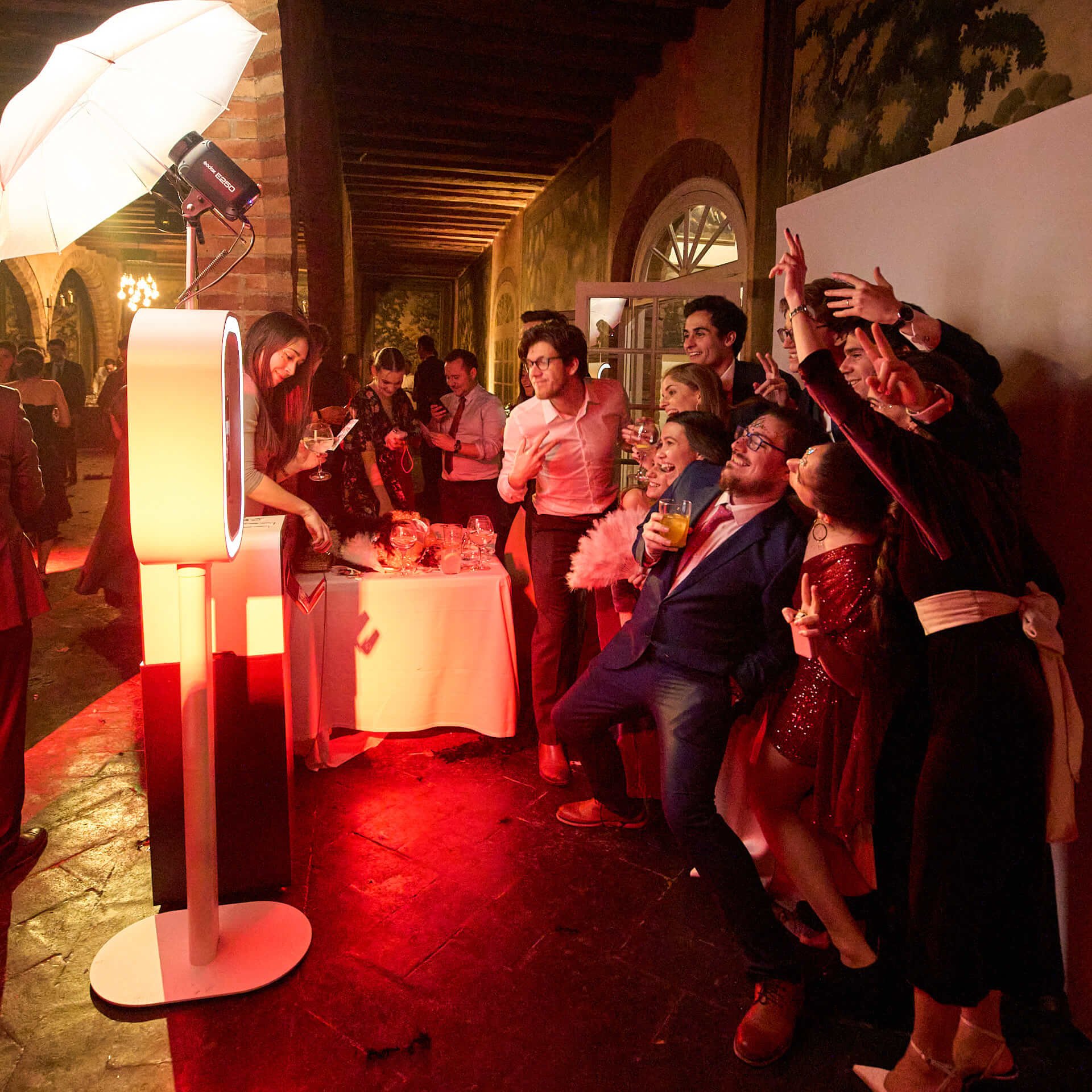 Group of people at a party using a photo booth with a ring light, some holding drinks, enjoying themselves with smiling and making peace signs.
