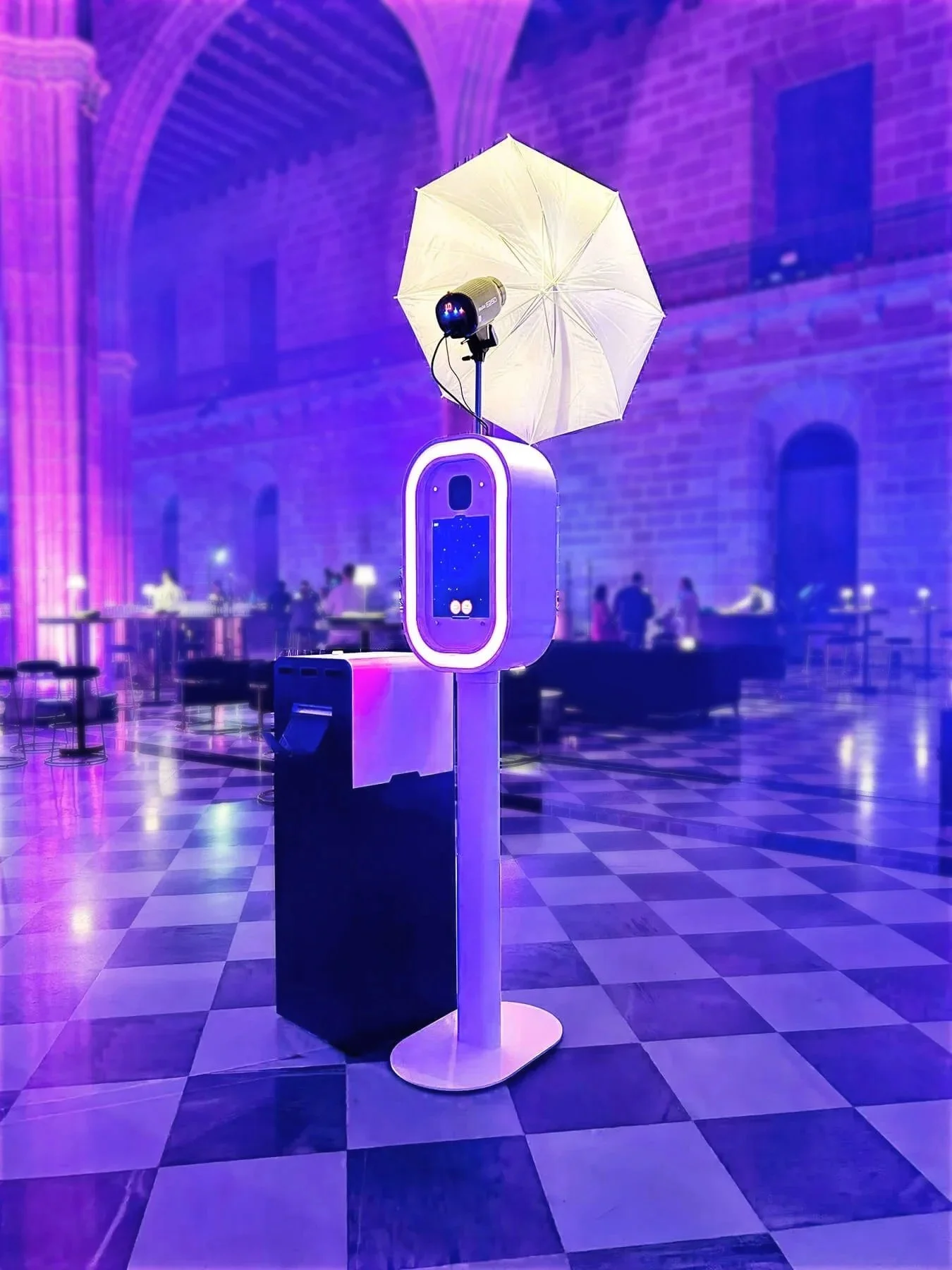 A photo booth station with a white umbrella, a speaker, and a screen displaying a starry sky, set in a room with purple lighting, arched ceilings, and checkered floor tiles.