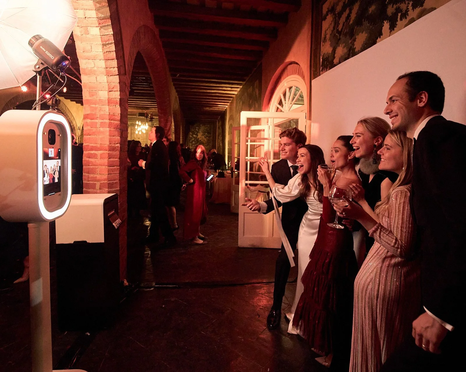 Group of six friends taking a selfie at a party, smiling and holding drinks, with a photo booth on the left.