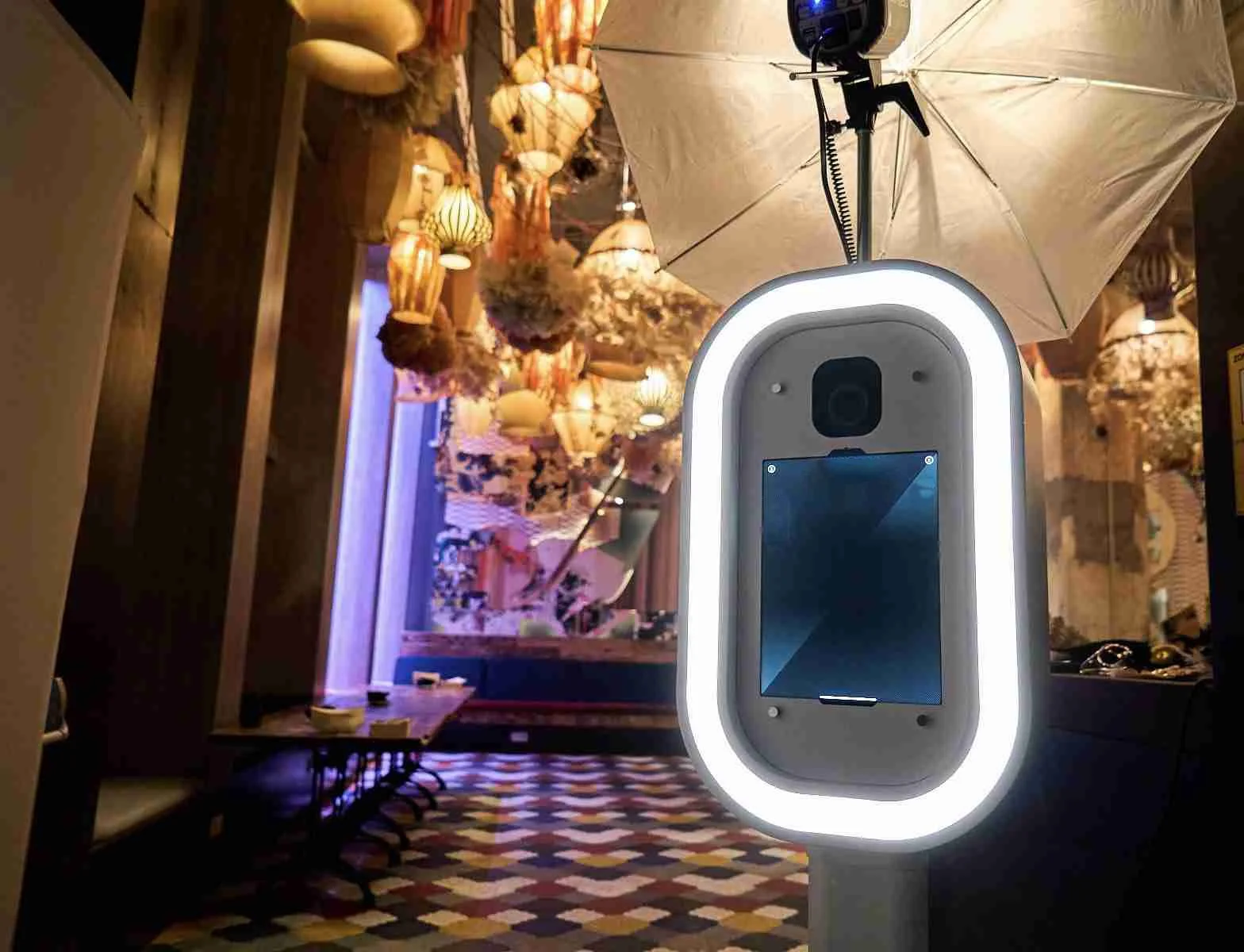 A photo booth with a large illuminated ring light, umbrella, and camera in a restaurant or café decorated with hanging lanterns and dried flowers.