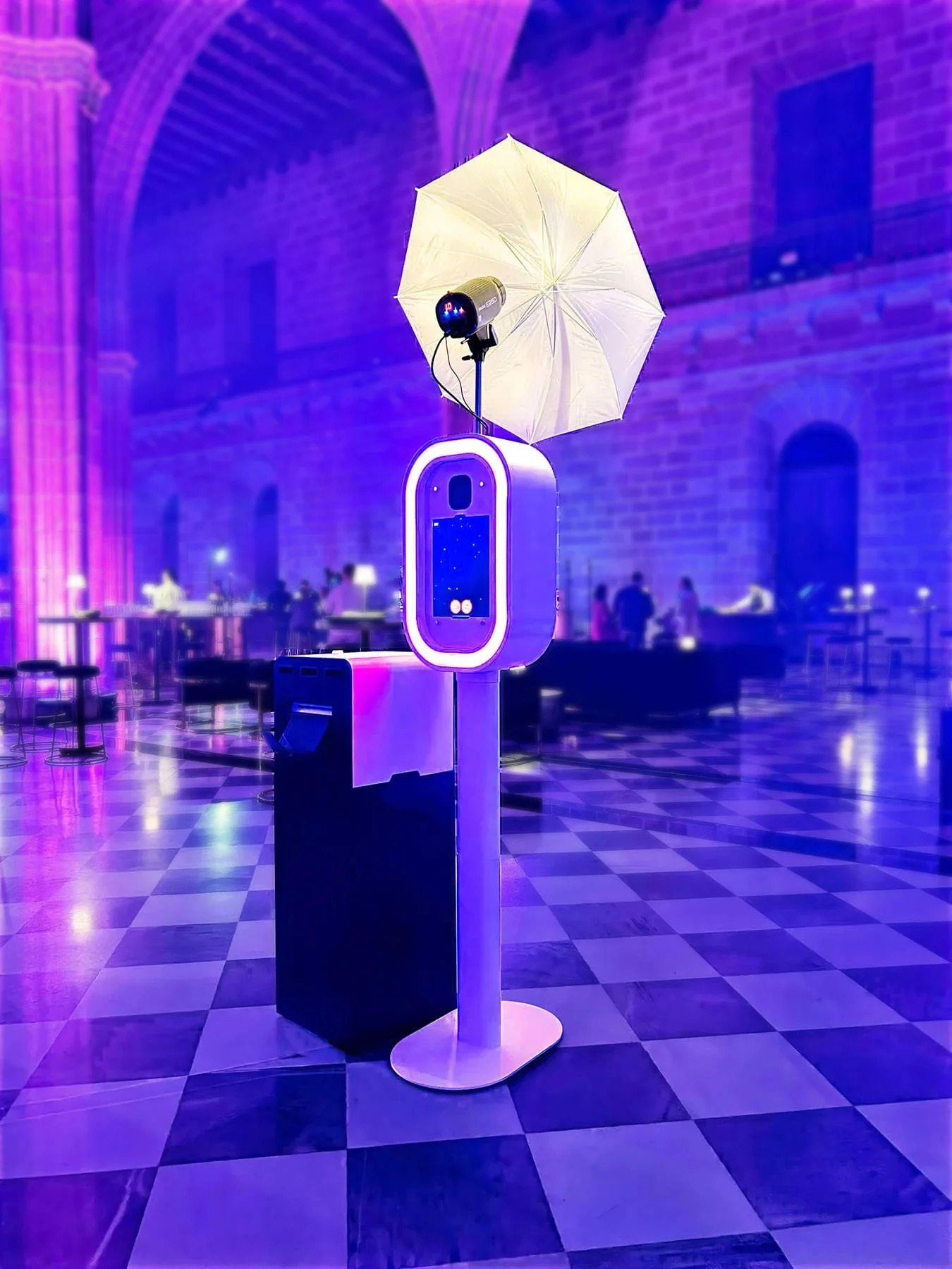 A photo of a photobooth with an umbrella attached, set in a brightly lit indoor space with checkered floor and purple lighting.