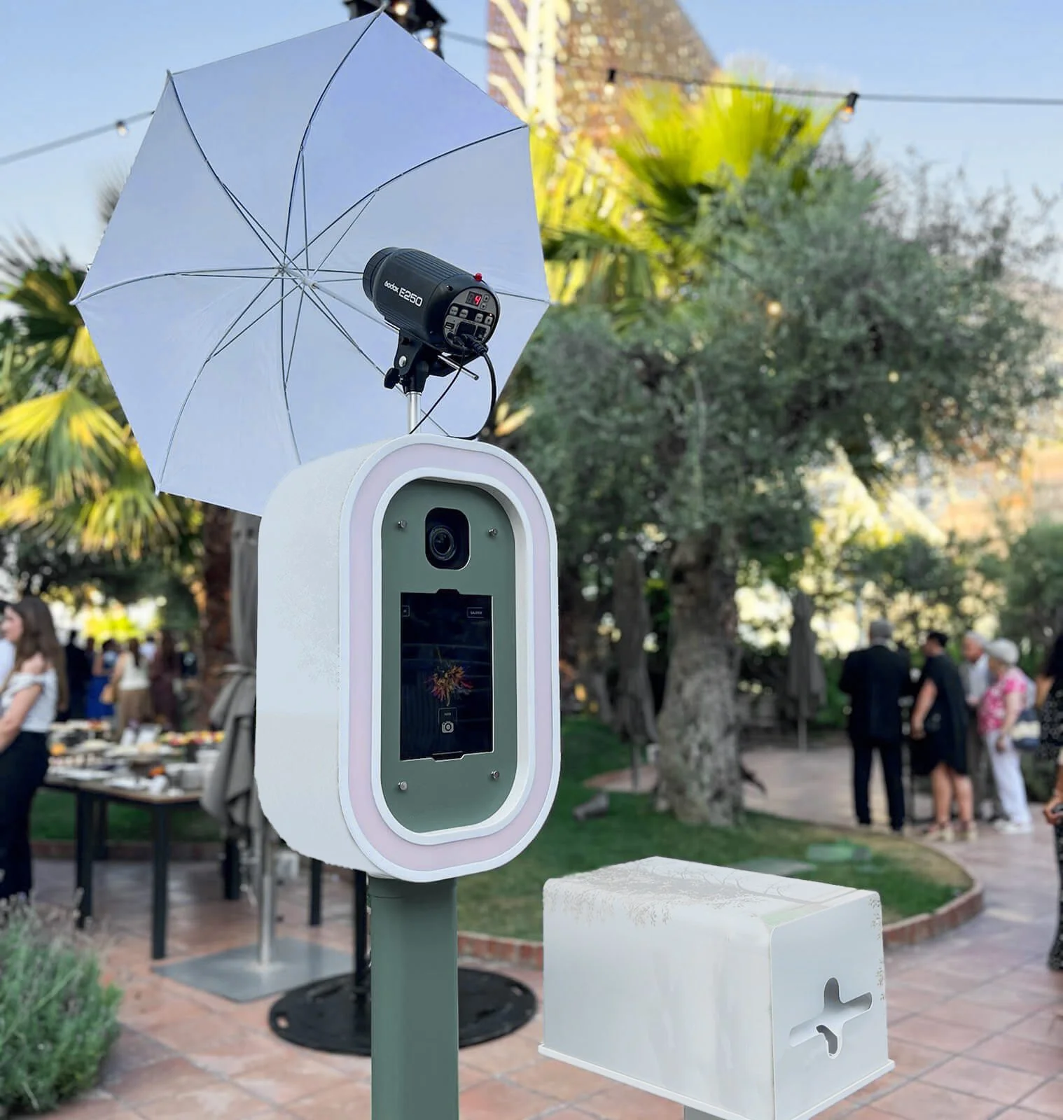 Self-service photo booth with a camera, touchscreen, an umbrella, and a light on a pole at an outdoor event with people in the background.
