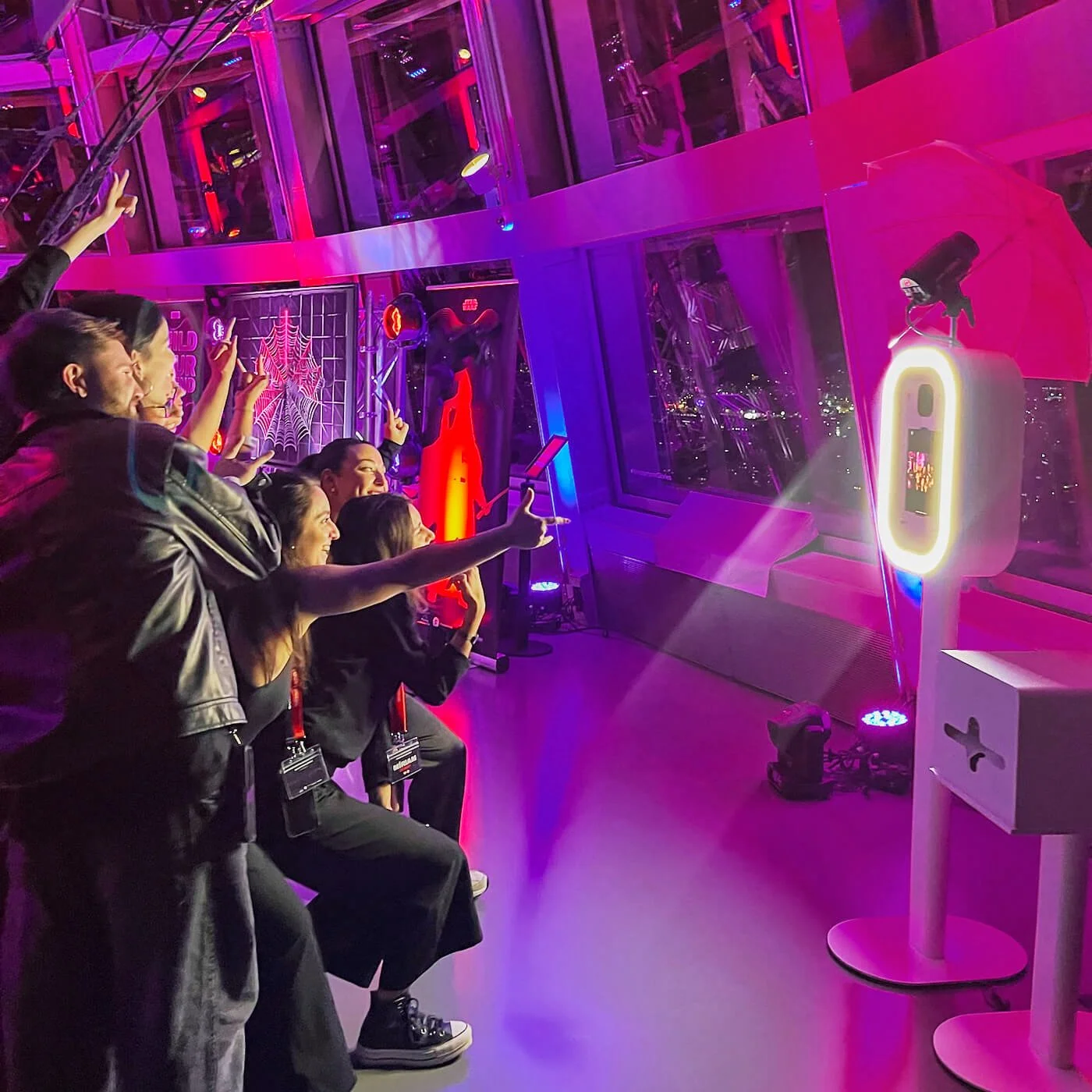 Group of people taking photos of neon art and spider web decorations on a starry night view through large windows.