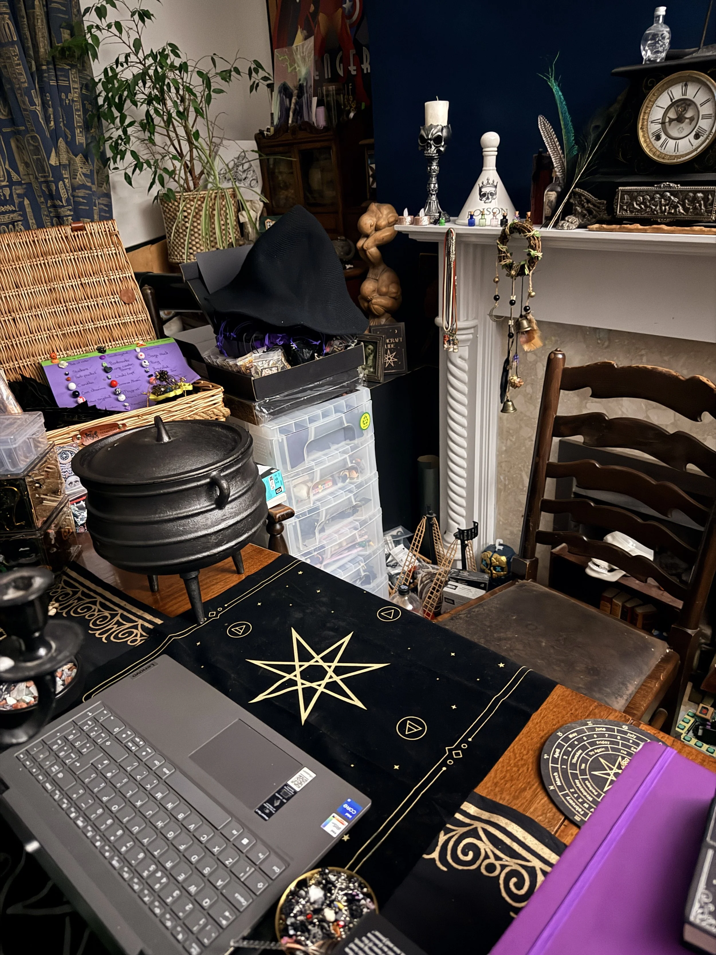 A cluttered room with a wooden table covered by a black and gold occult-themed tablecloth, a laptop, and various mystical and decorative items. To the left, there is a basket with jewelry, and a black cauldron. In the background, there are storage containers, plants, and a white fireplace mantel decorated with candles, jewelry, and skull ornaments. A clock and a bottle are on top of the mantel, and there are chairs around the table.