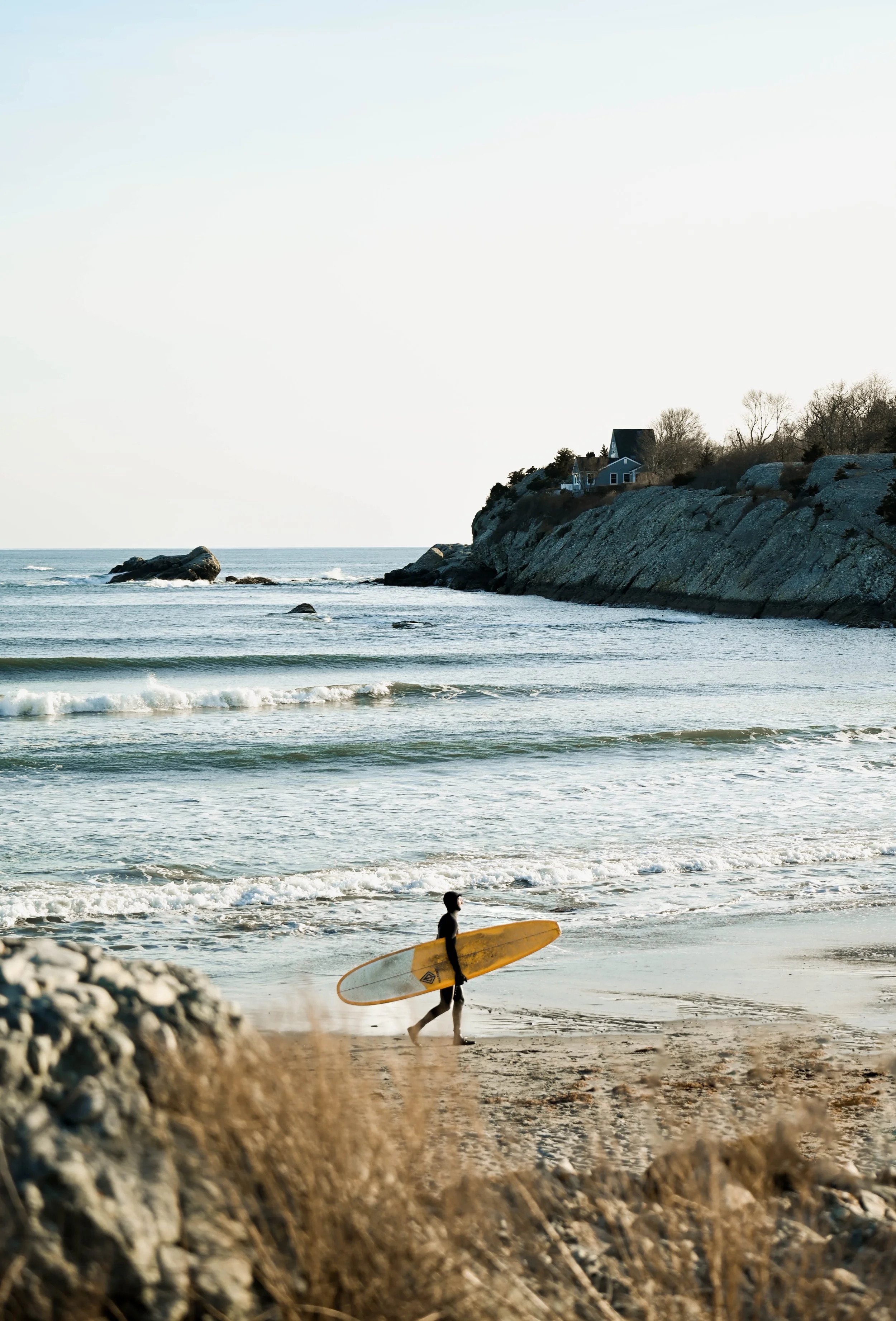 A person walking along a beach carrying a surfboard with rocks, ocean waves, and a house on a cliff in the background.