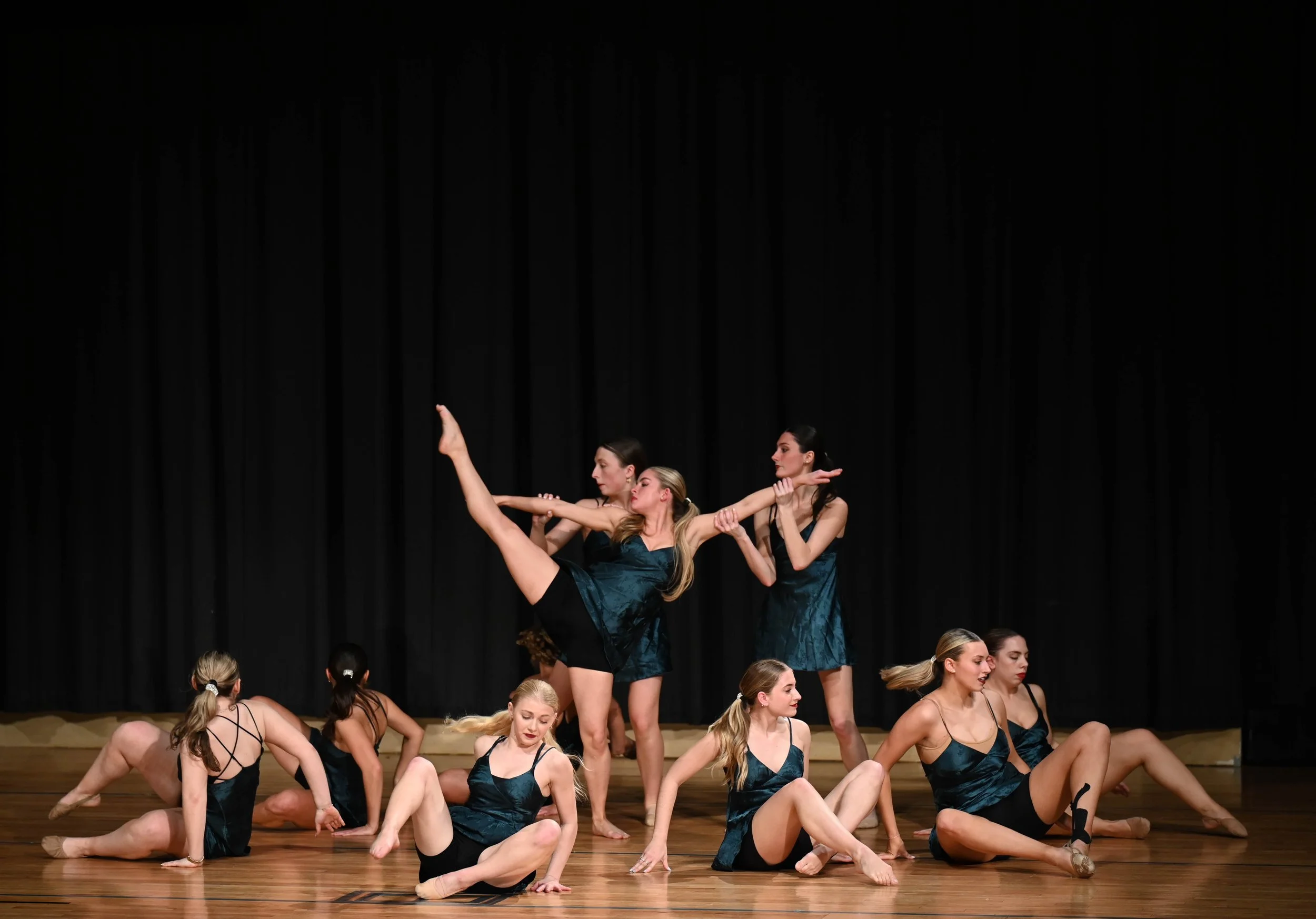 Group of young women performing a synchronized dance routine on stage, with one dancer lifting a leg and others in various poses, all wearing matching dark teal dresses against a black curtain background.