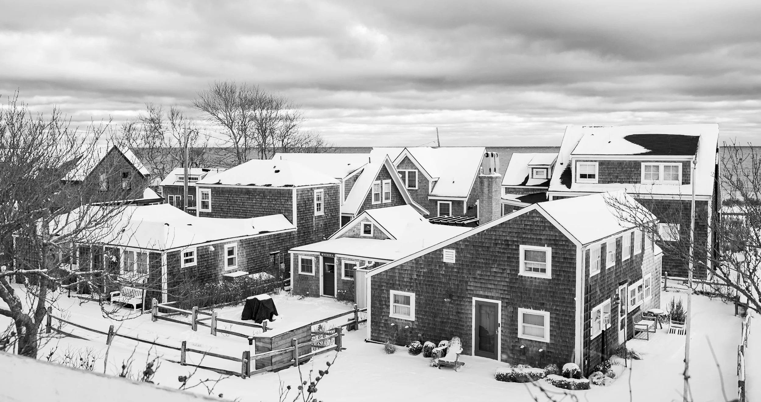 Snow-covered houses in a residential neighborhood during winter, with bare trees and cloudy sky.