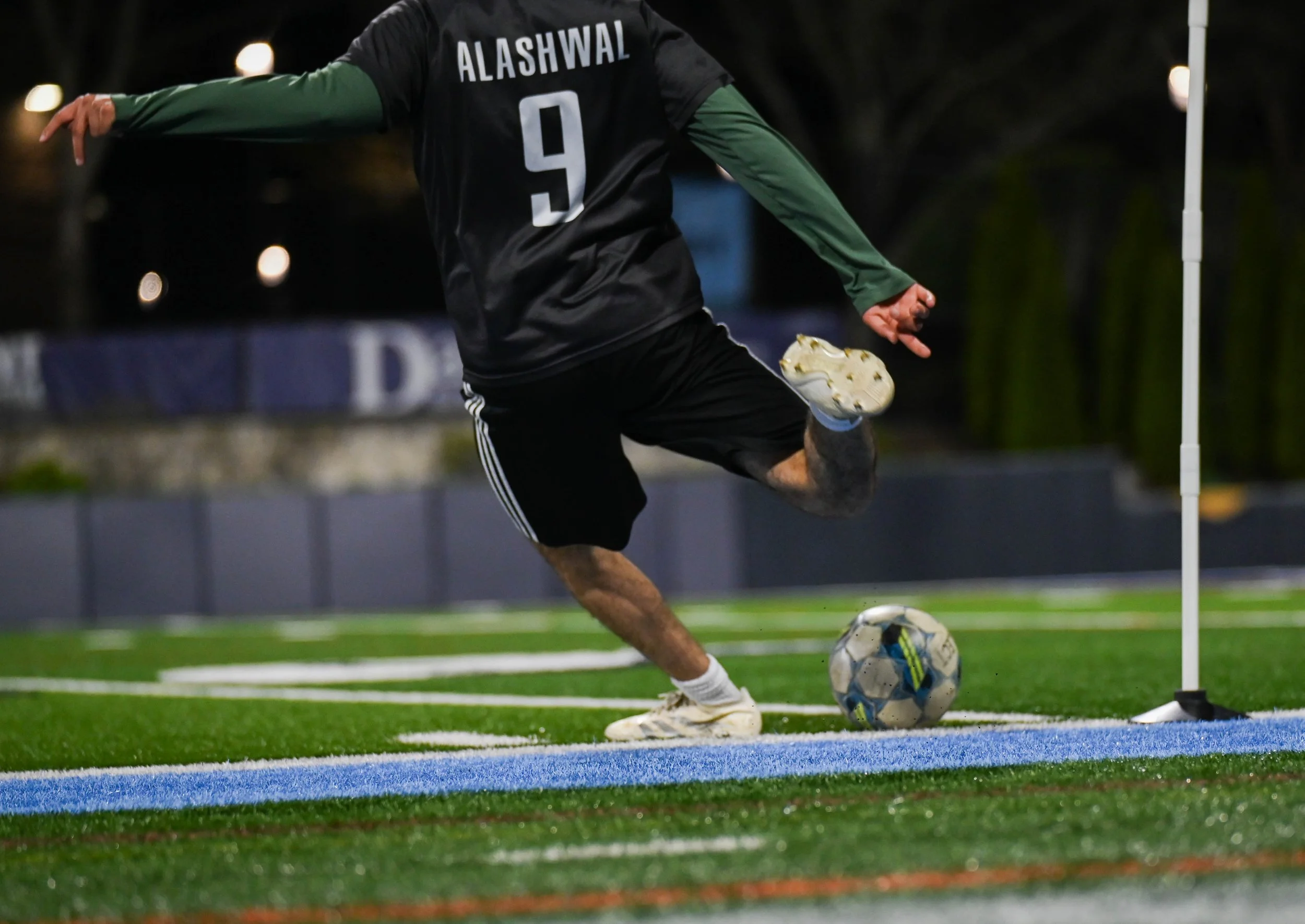 A soccer player wearing a black jersey with the name Alashwal and number 9 is kicking a soccer ball on a field during a match at night.
