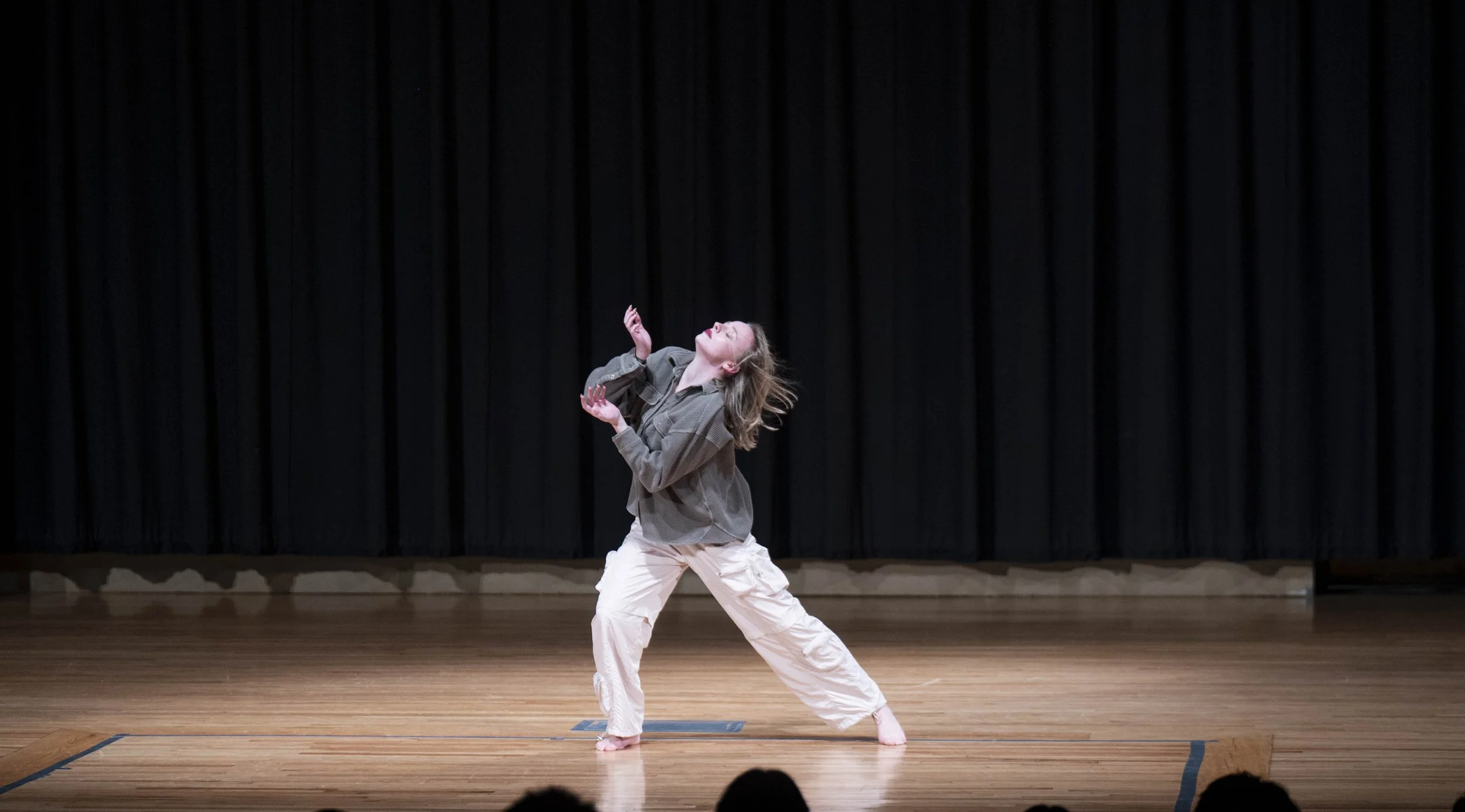 A woman dancing on stage with a black curtain background, wearing a gray jacket and white pants.