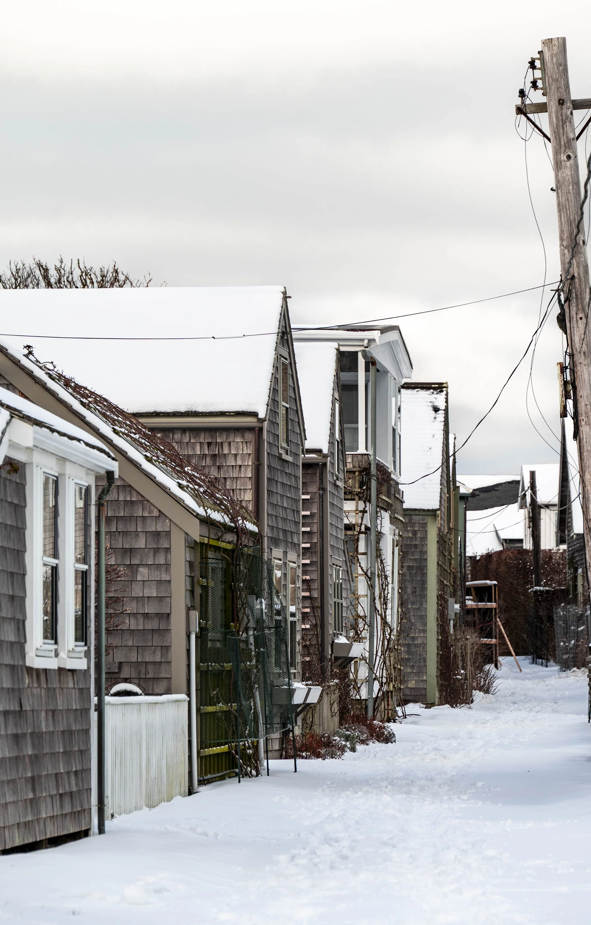 Snow-covered alleyway lined with weathered wooden and shingle-clad houses, with utility poles and power lines against a cloudy sky.
