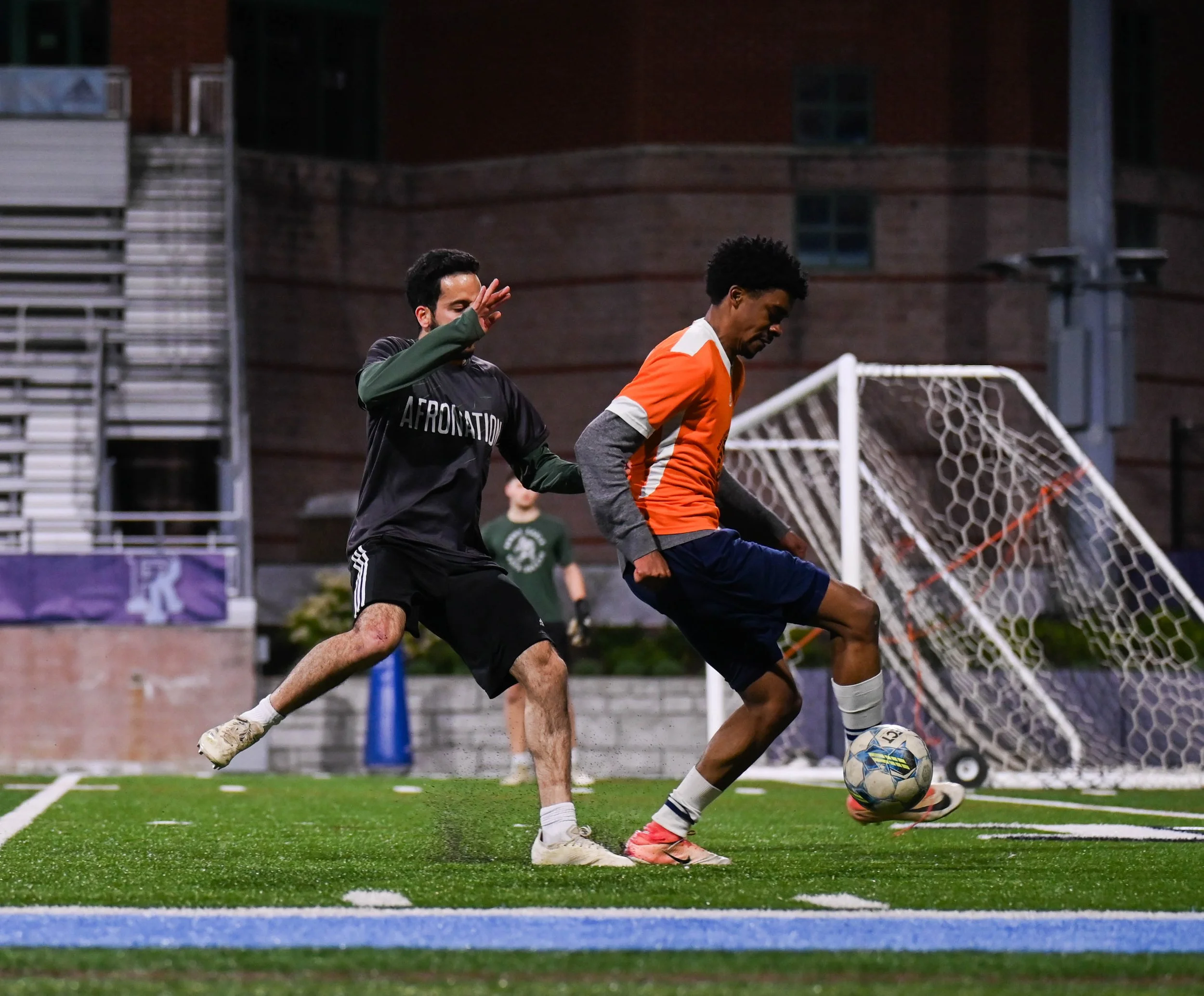 Two soccer players compete for the ball on an outdoor field at night, with a goal net in the background.