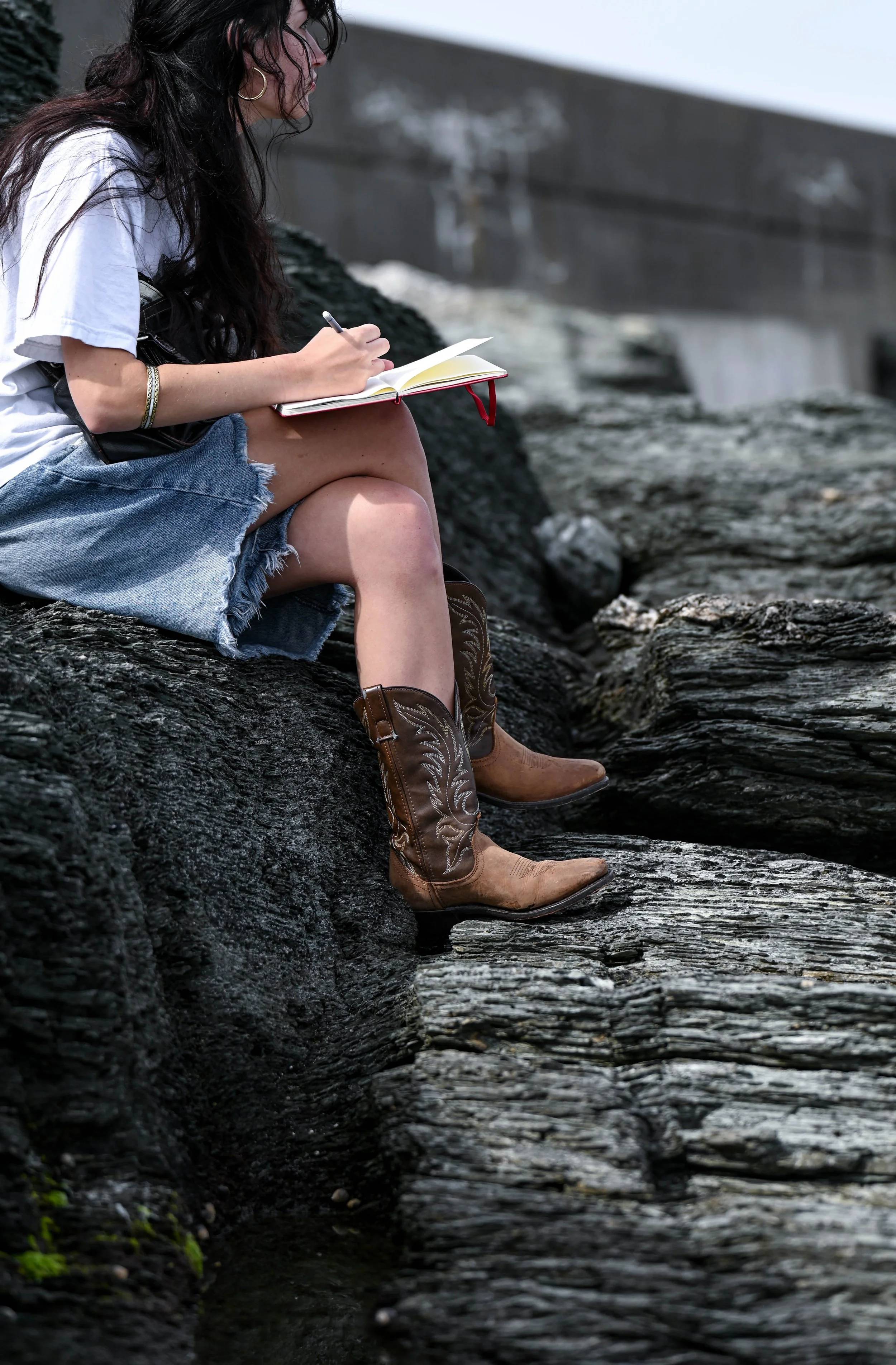 A woman sitting on rocks near water, writing in a notebook while wearing a white t-shirt, denim shorts, and cowboy boots.