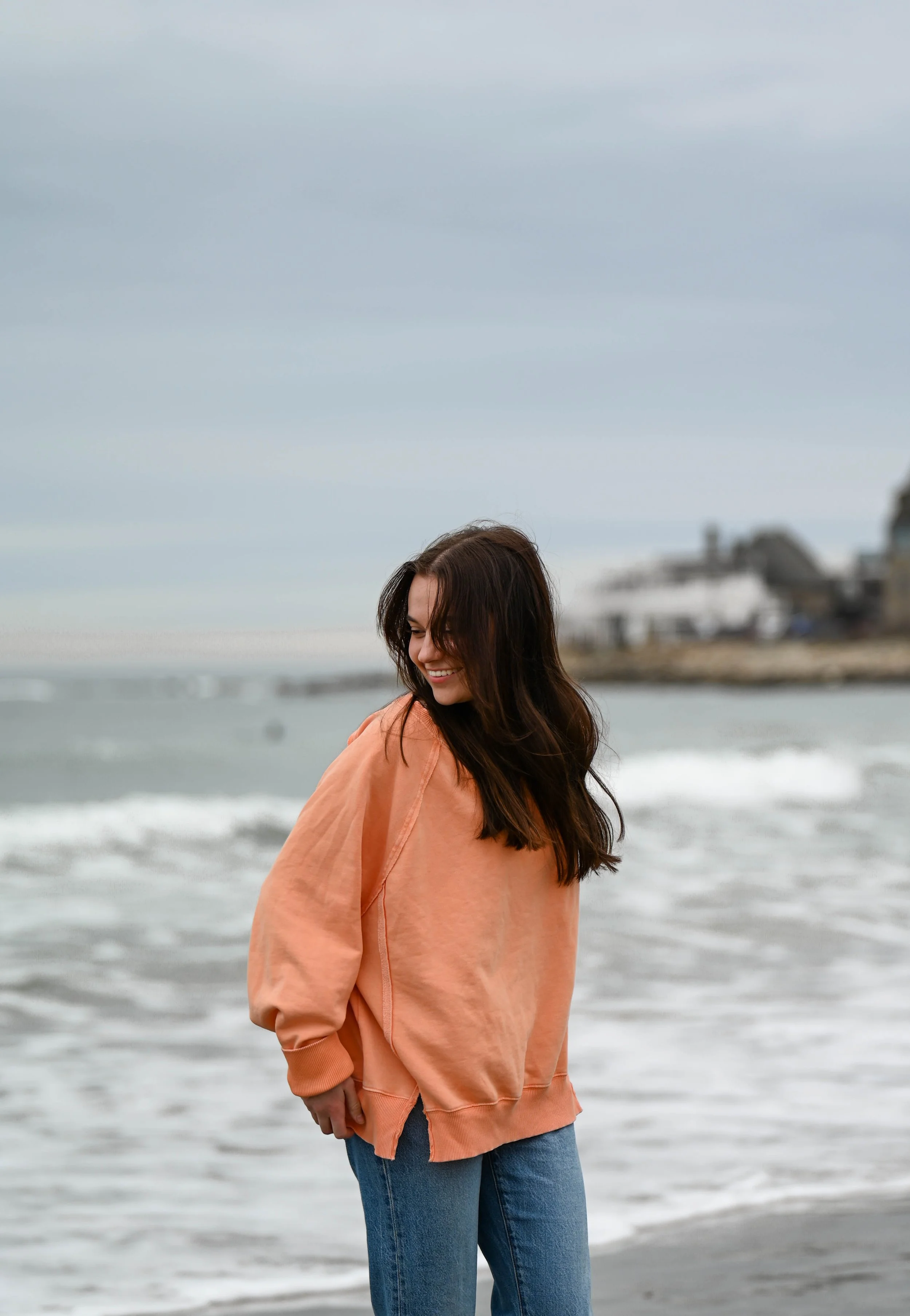 A young woman with long dark hair, wearing an orange sweatshirt and blue jeans, standing on the beach near the water, smiling and looking downward, overcast sky in the background.