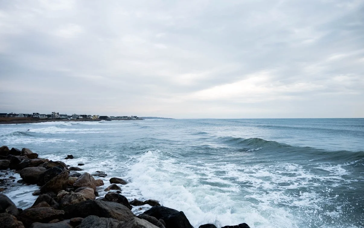 Overcast view of the ocean with waves crashing against rocky shoreline and houses in the distance.