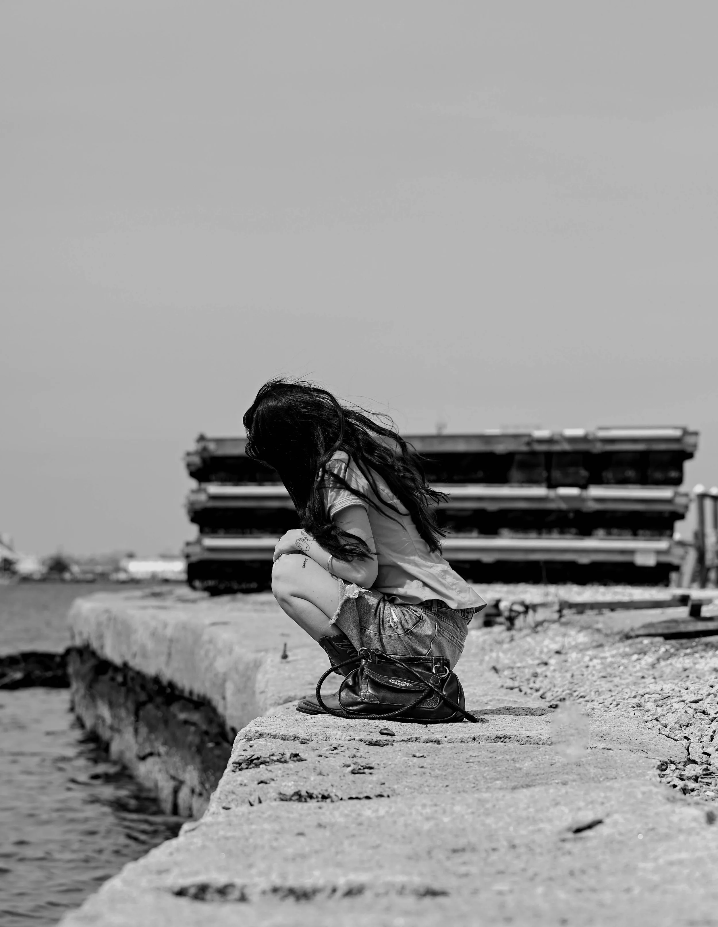 A woman with long dark hair squats on a concrete dock by the water, facing away from the camera, with stacked containers in the background.