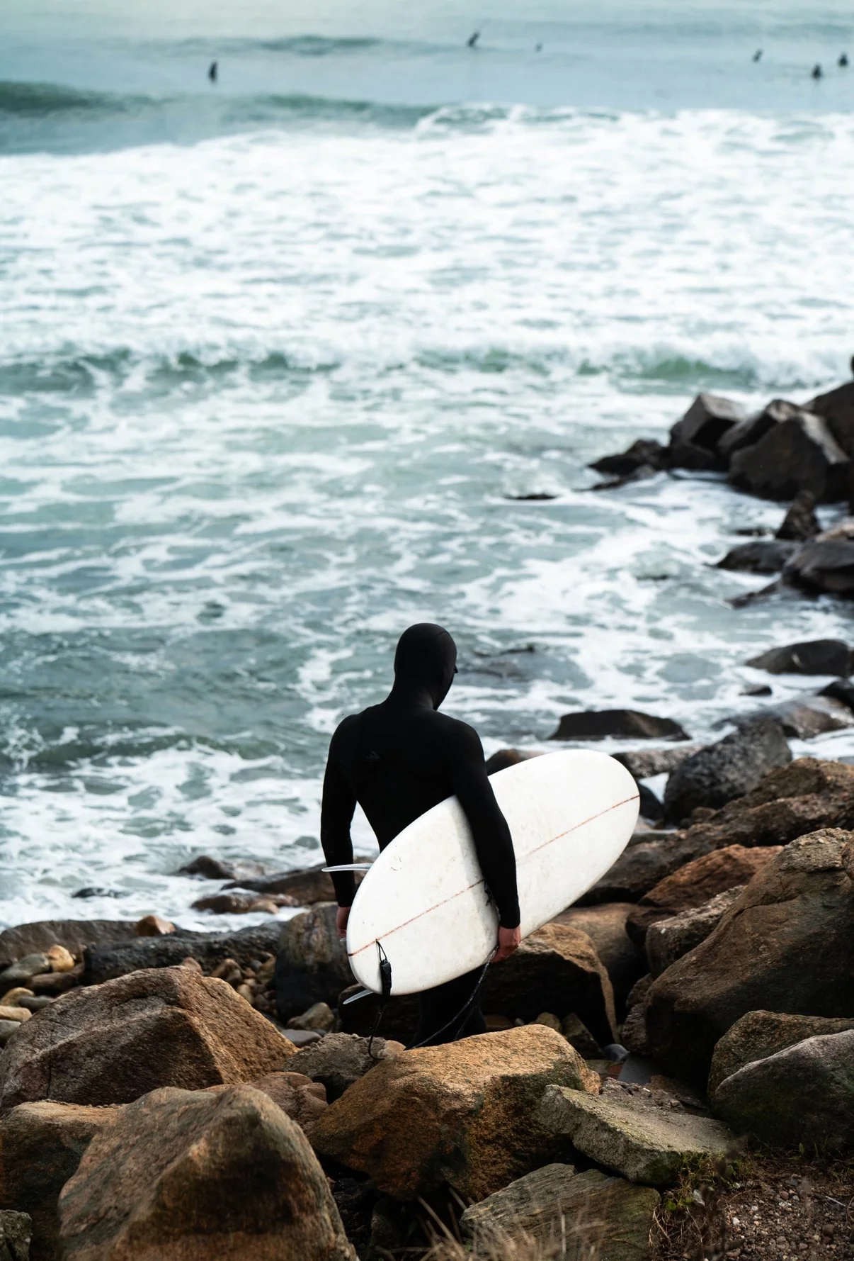 A person wearing a black wetsuit holding a white surfboard, standing on a rocky shore with waves in the background.