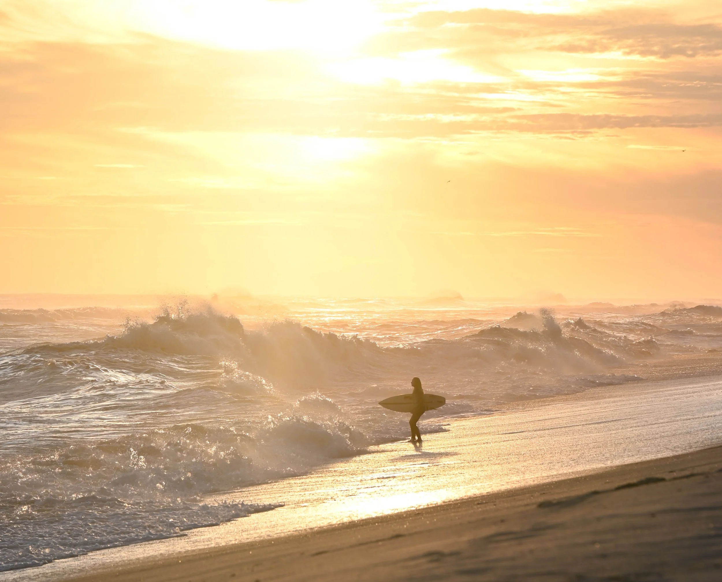 A surfer walking along the beach at sunset carrying a surfboard with waves crashing behind.