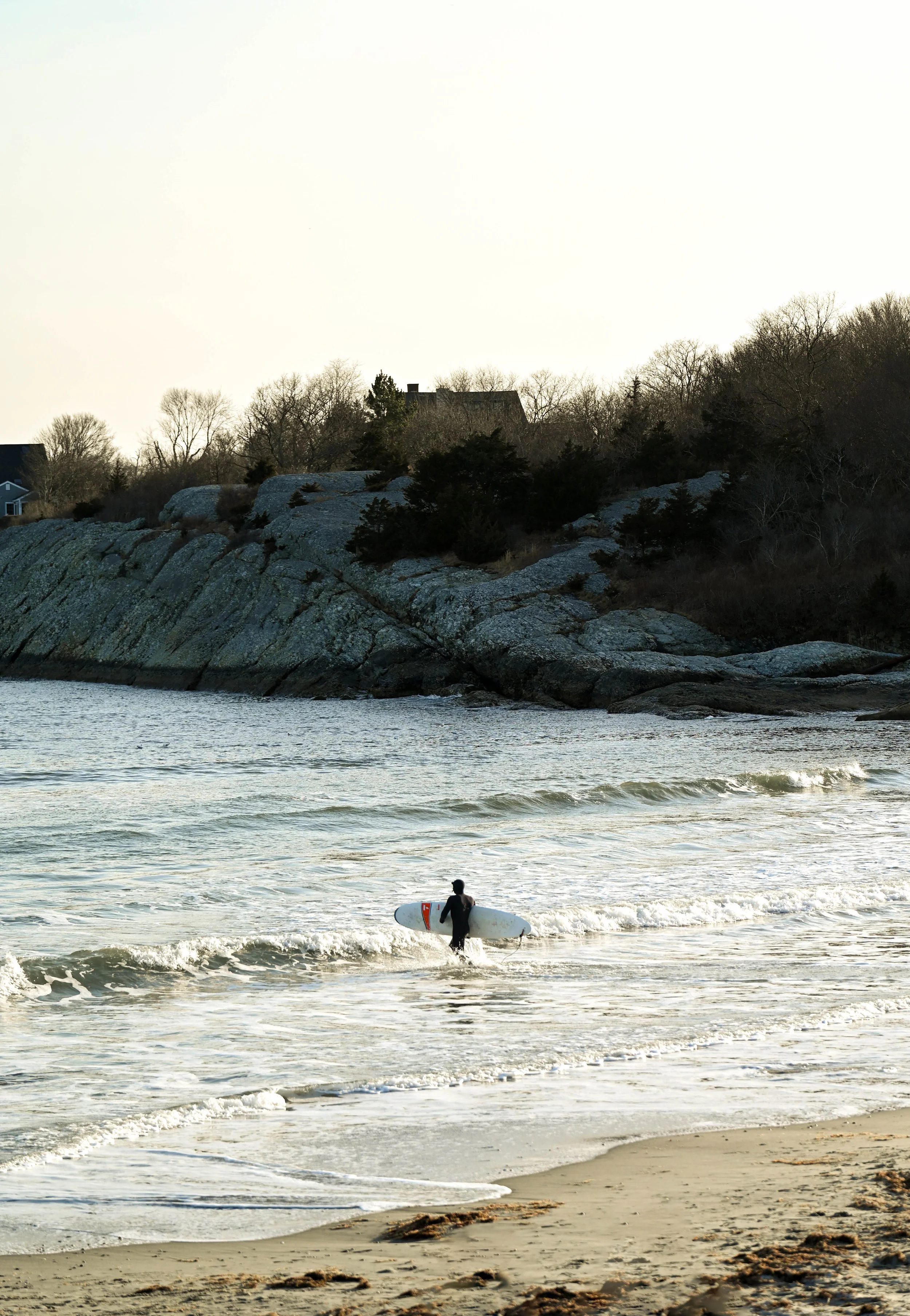 A person holding a surfboard walking into the water at a sandy beach with rocky cliffs and trees in the background.
