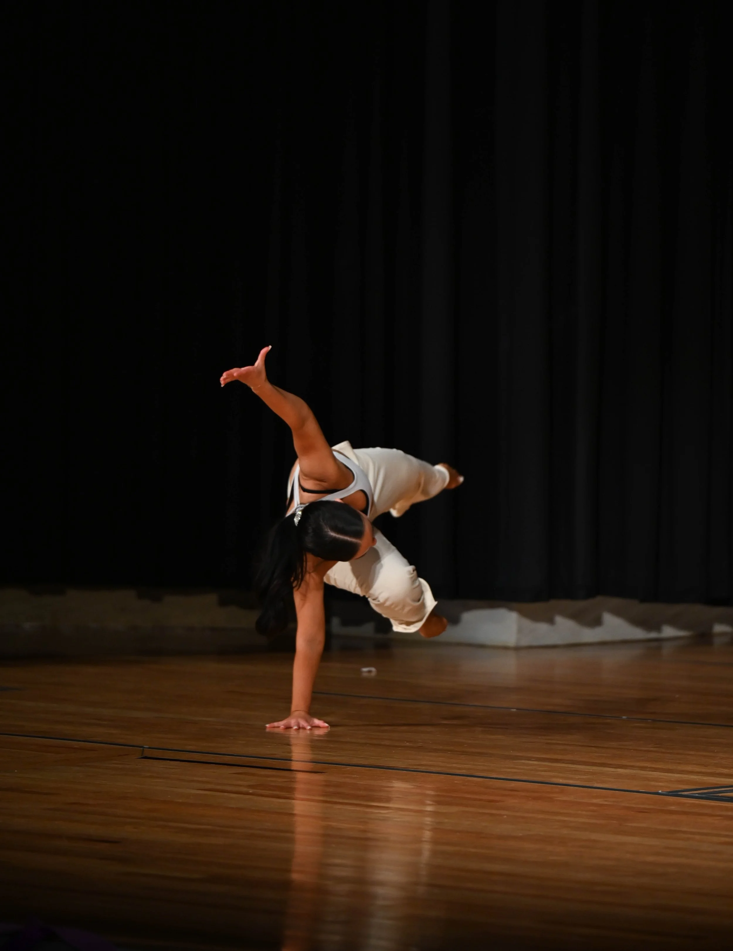 A woman performing a breakdance move on a wooden stage with black curtains in the background.