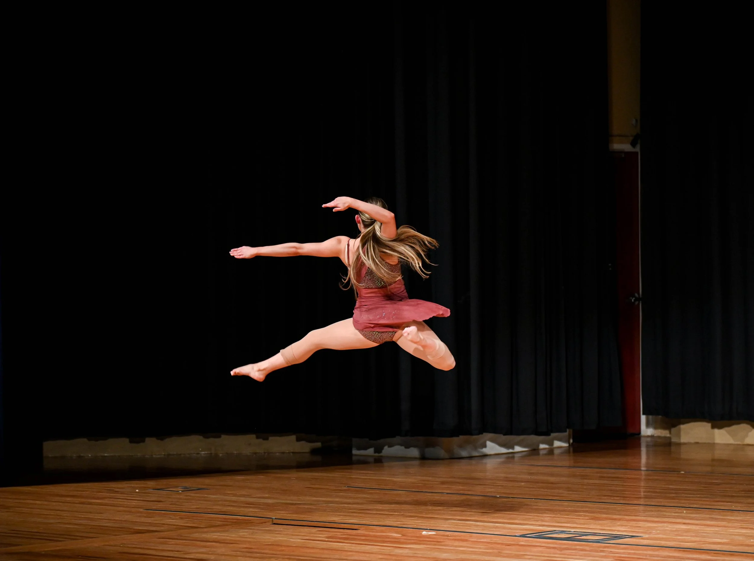A female dancer mid-air jump on a wooden stage with black curtains in the background.