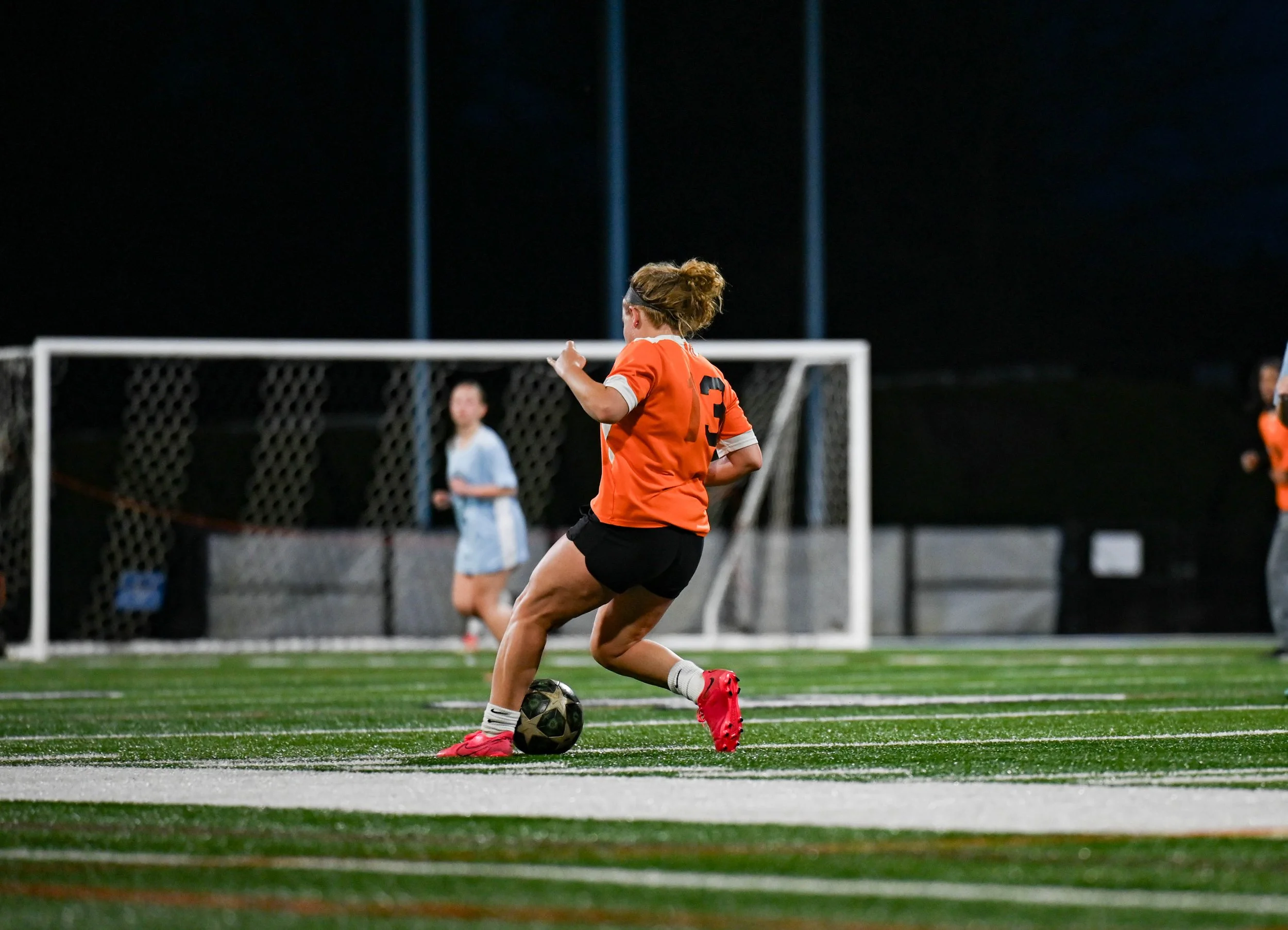 Female soccer player in orange jersey and black shorts kicking a ball during a nighttime game on a synthetic turf field.