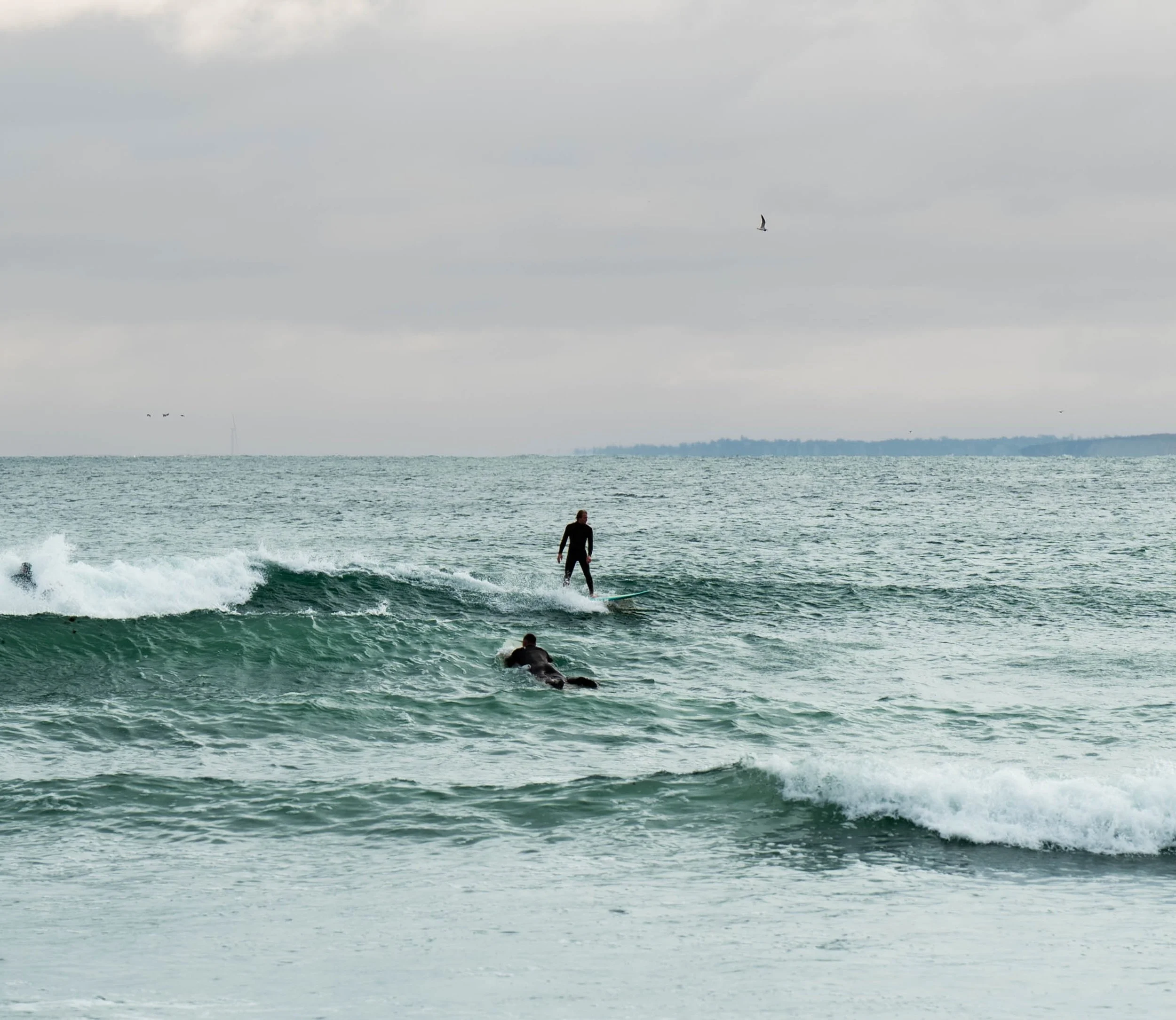 Two surfers in wetsuits on a cloudy day, one riding a wave and the other paddling in the water, with an ocean and distant shoreline in the background.