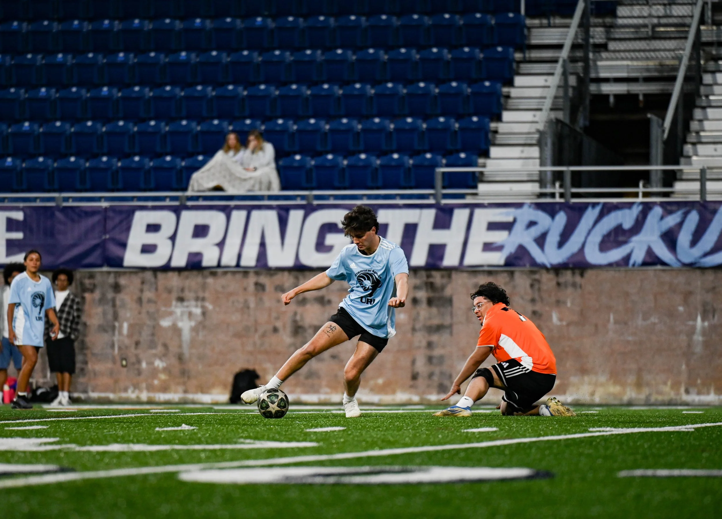 A soccer player in a light blue jersey and black shorts kicking a soccer ball towards the goal on a green turf field. Another player in an orange jersey is kneeling on the ground nearby. In the background, a banner with the text 'BRING THE RUCKUS' an