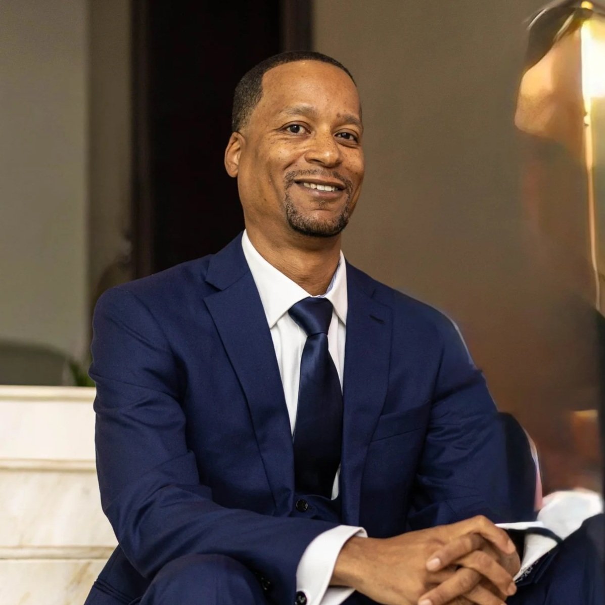 A man in a navy blue suit and tie smiling, sitting with hands clasped, in an indoor setting.