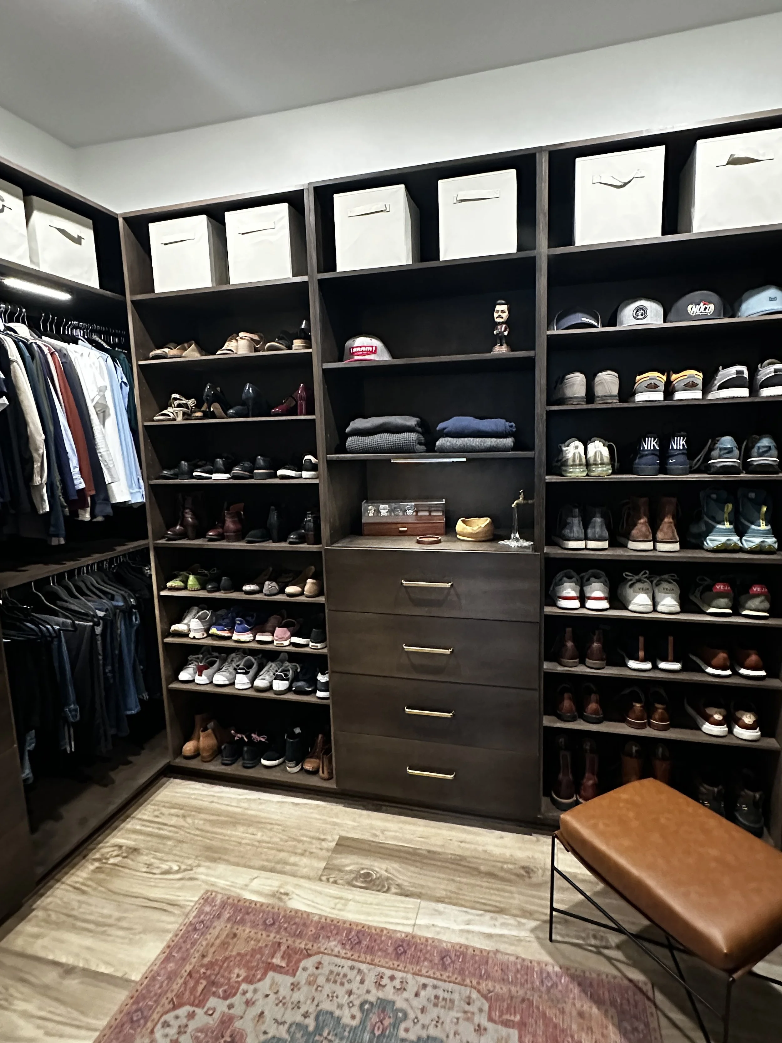 A walk-in closet with dark wooden shelving filled with shoes, hats, clothes, and storage boxes. There is a small cushioned bench in the foreground over a patterned rug.