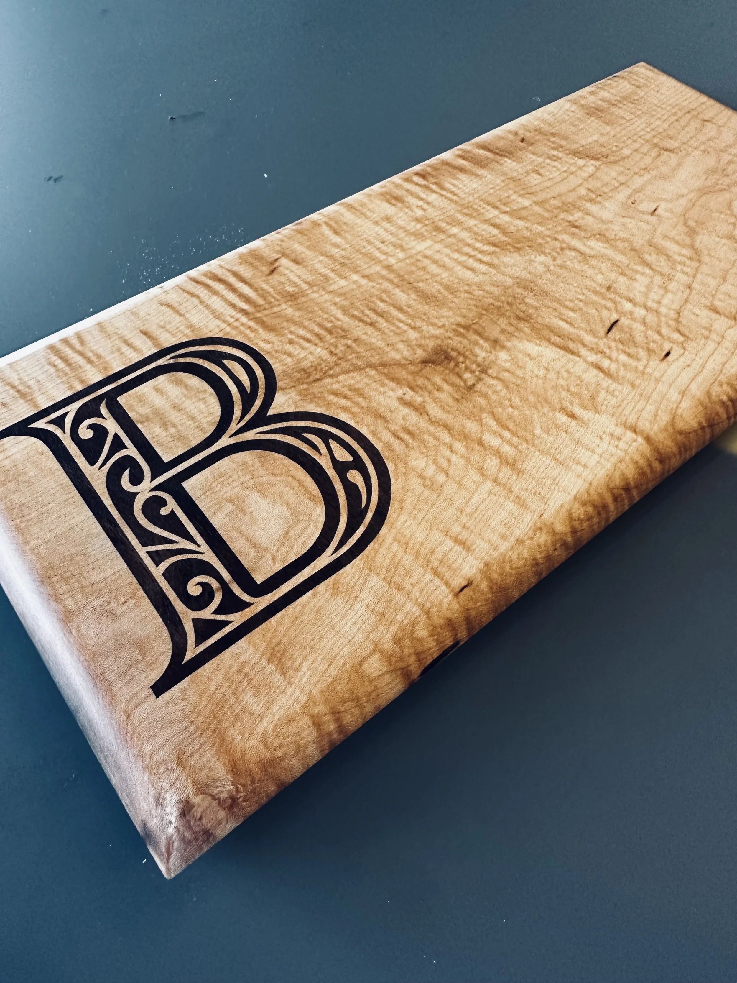 Wooden cutting board with a decorative black letter B printed on it, placed on a dark surface.