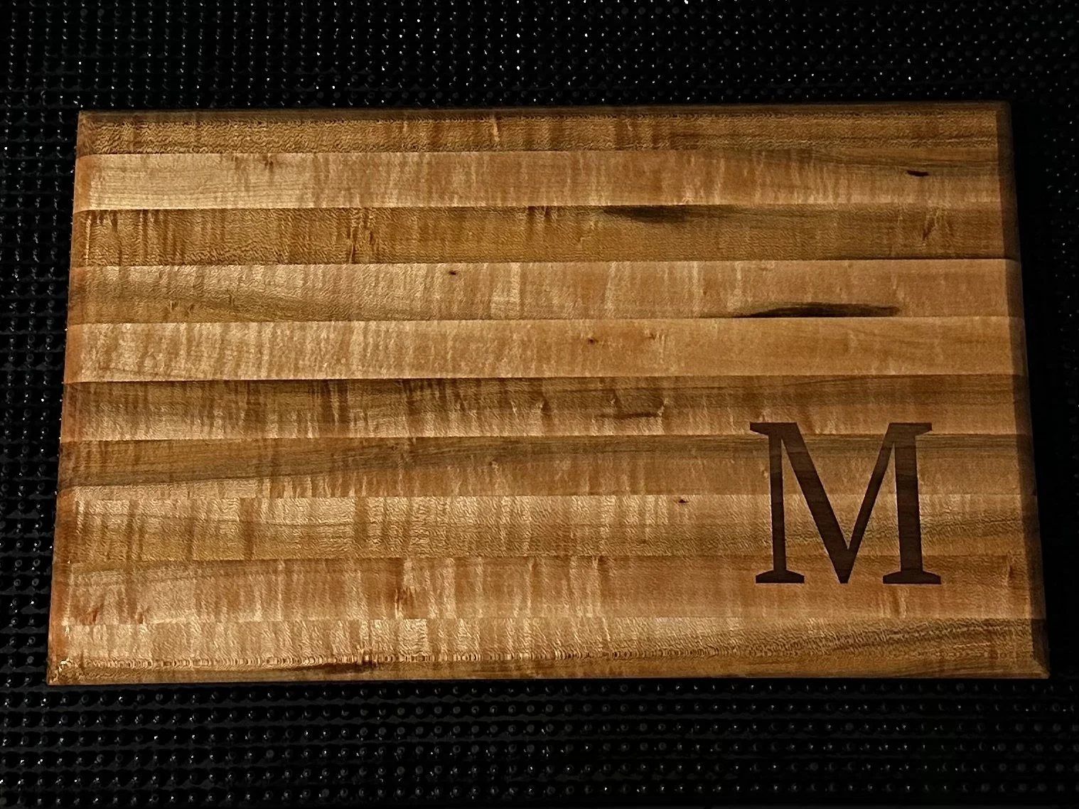 A wooden cutting board with the letter 'M' engraved in the bottom right corner, placed on a black textured surface.
