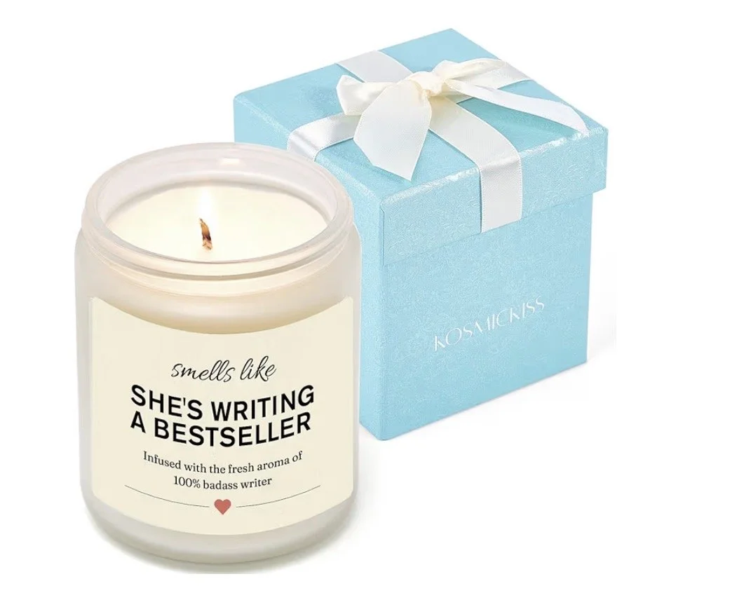 A white candle with a label that says 'smells like she's writing a bestseller' and a small red heart. The candle is in front of a light blue gift box with a white ribbon.