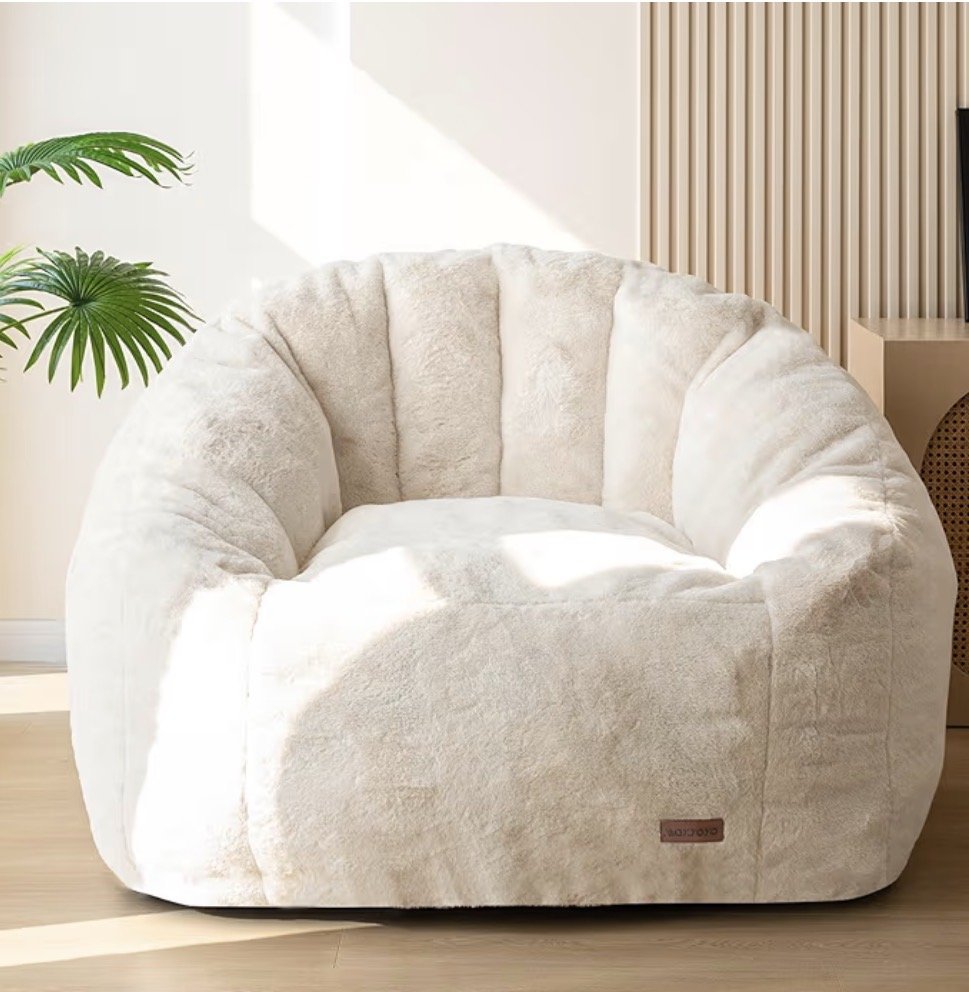A plush, cream-colored, rounded armchair or lounge chair in a bright, modern room with a green plant on the left and a wooden slat wall on the right.