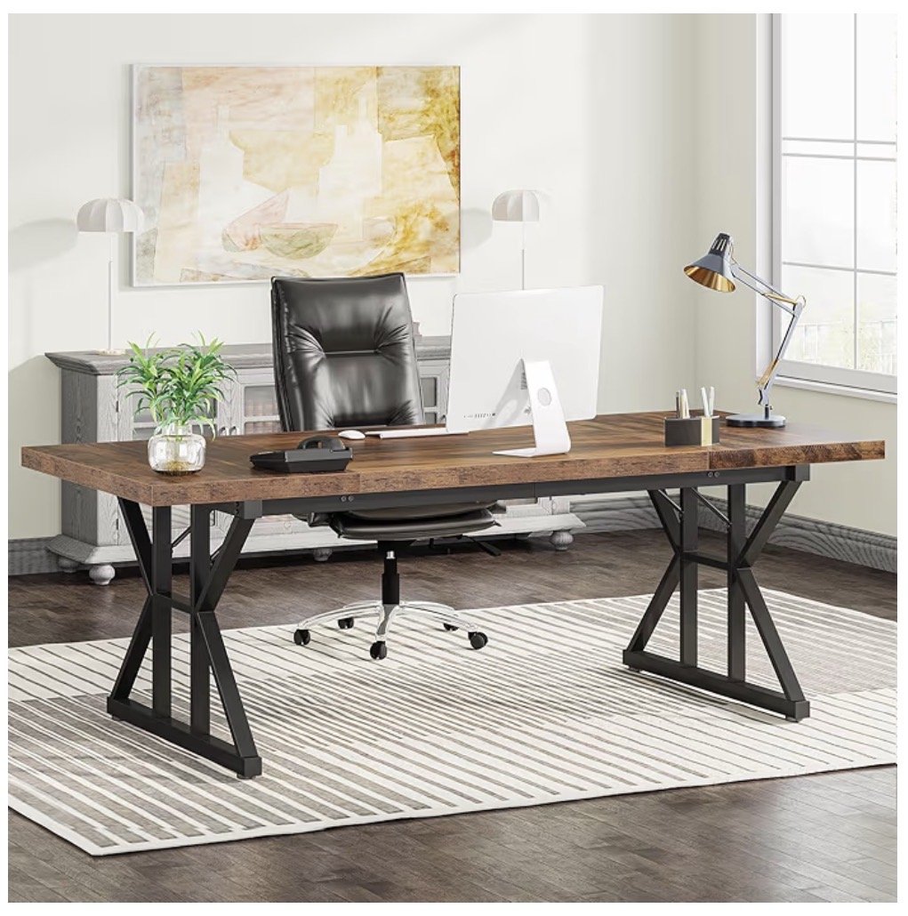 Modern office space with a large wooden desk, black leather chair, white desktop monitor, desk lamp, potted plant, and decorative items, with a painting on the wall and a window letting in natural light.