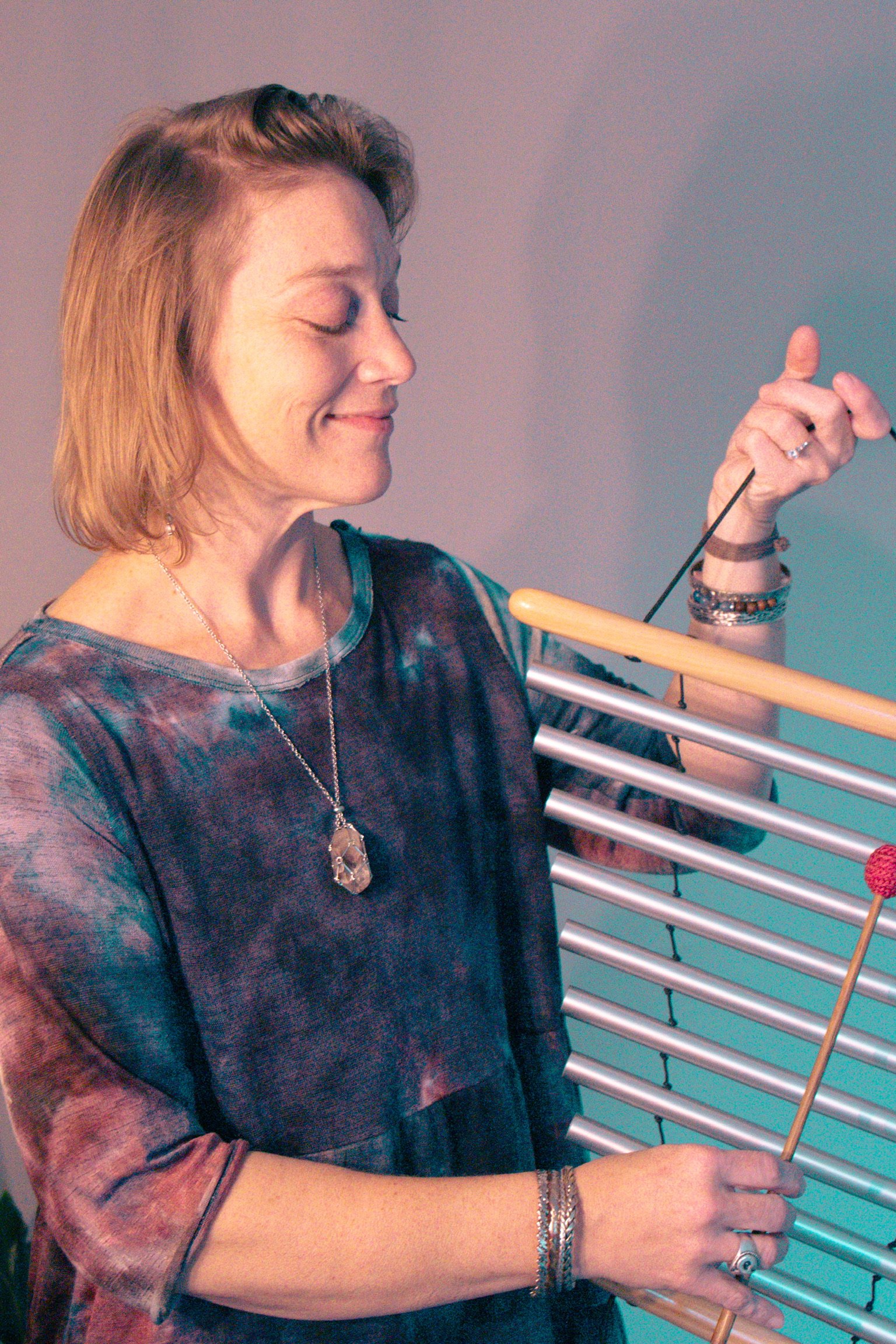 A woman with shoulder-length reddish brown hair playing a metal and wooden wind instrument with colorful mallets, wearing a multicolored tie-dye style shirt, jewelry, and a ring, smiling with her eyes closed.